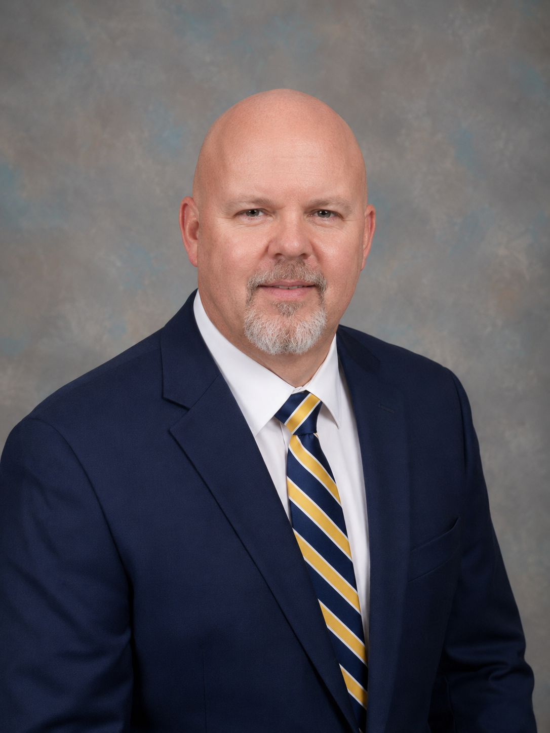 Man smiling at the camera in a navy suit, with a navy and yellow tie on a neutral background
