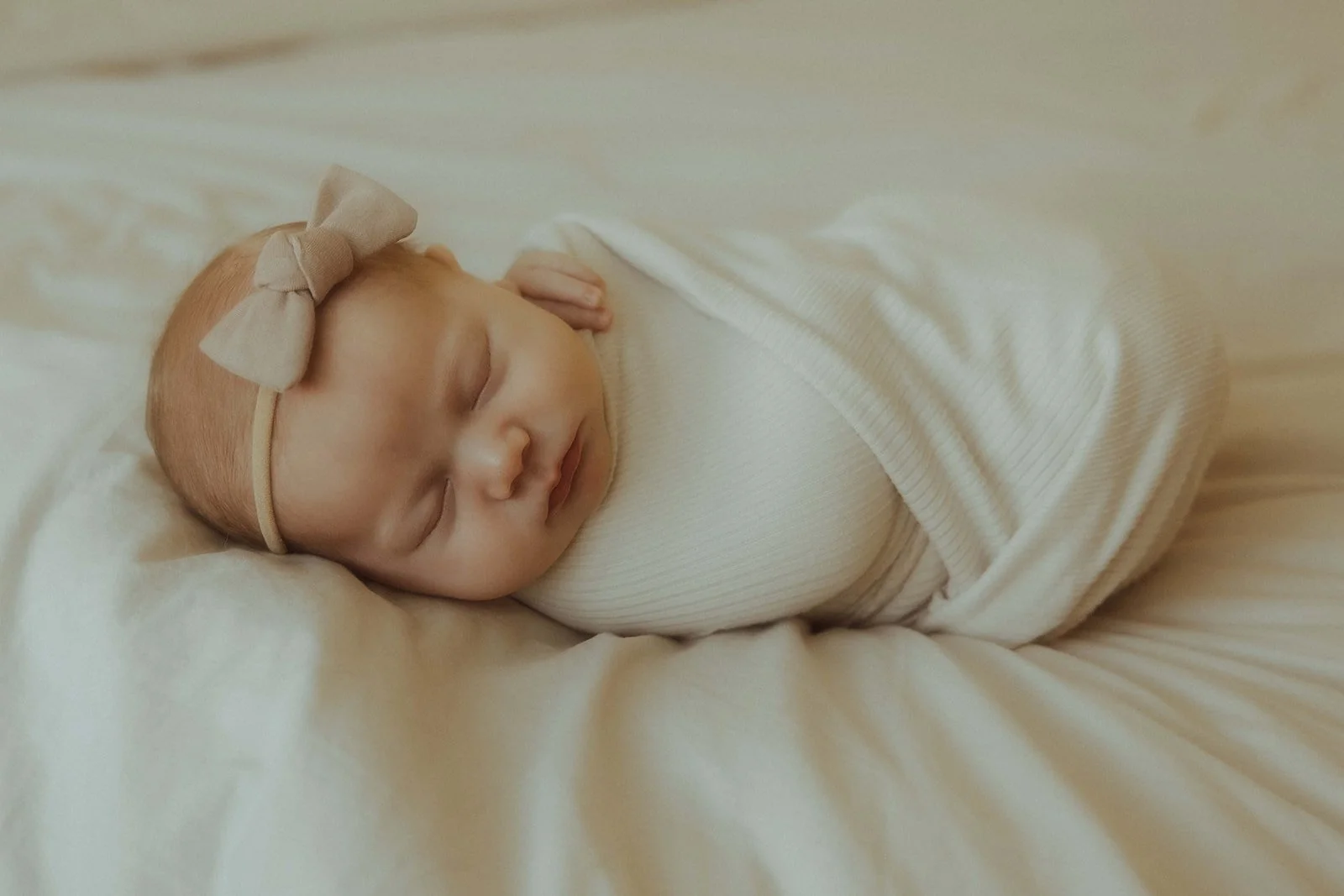 A newborn baby girl swaddled in a white swaddle while asleep and laying on a white bed.