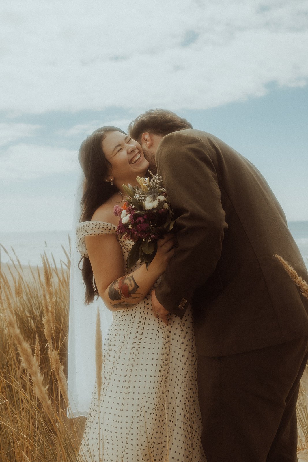 A bride and groom embracing on the beach while laughing, surrounded by beach, sand, and tall grass.