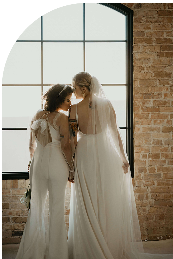 Two women on their wedding day standing near one another while one of them kisses the other's shoulder.