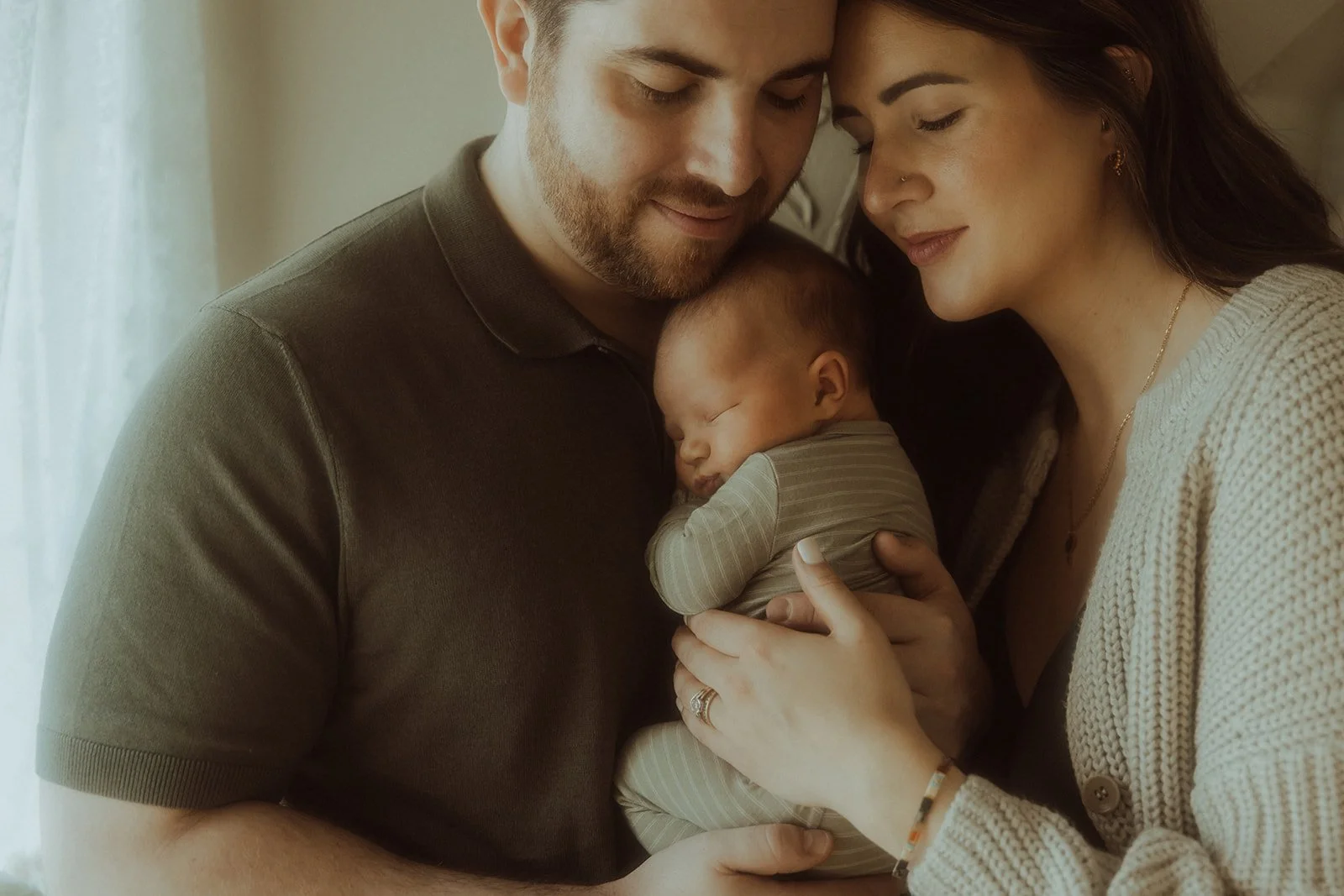 Two parents snuggling with their newborn baby boy near a window in a nursery.