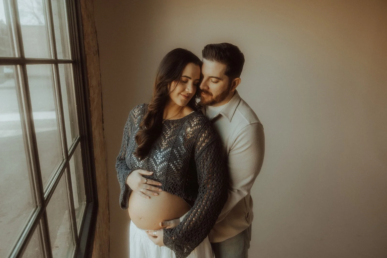 A couple embracing each other and cradling the women's pregnant stomach while standing near a window.