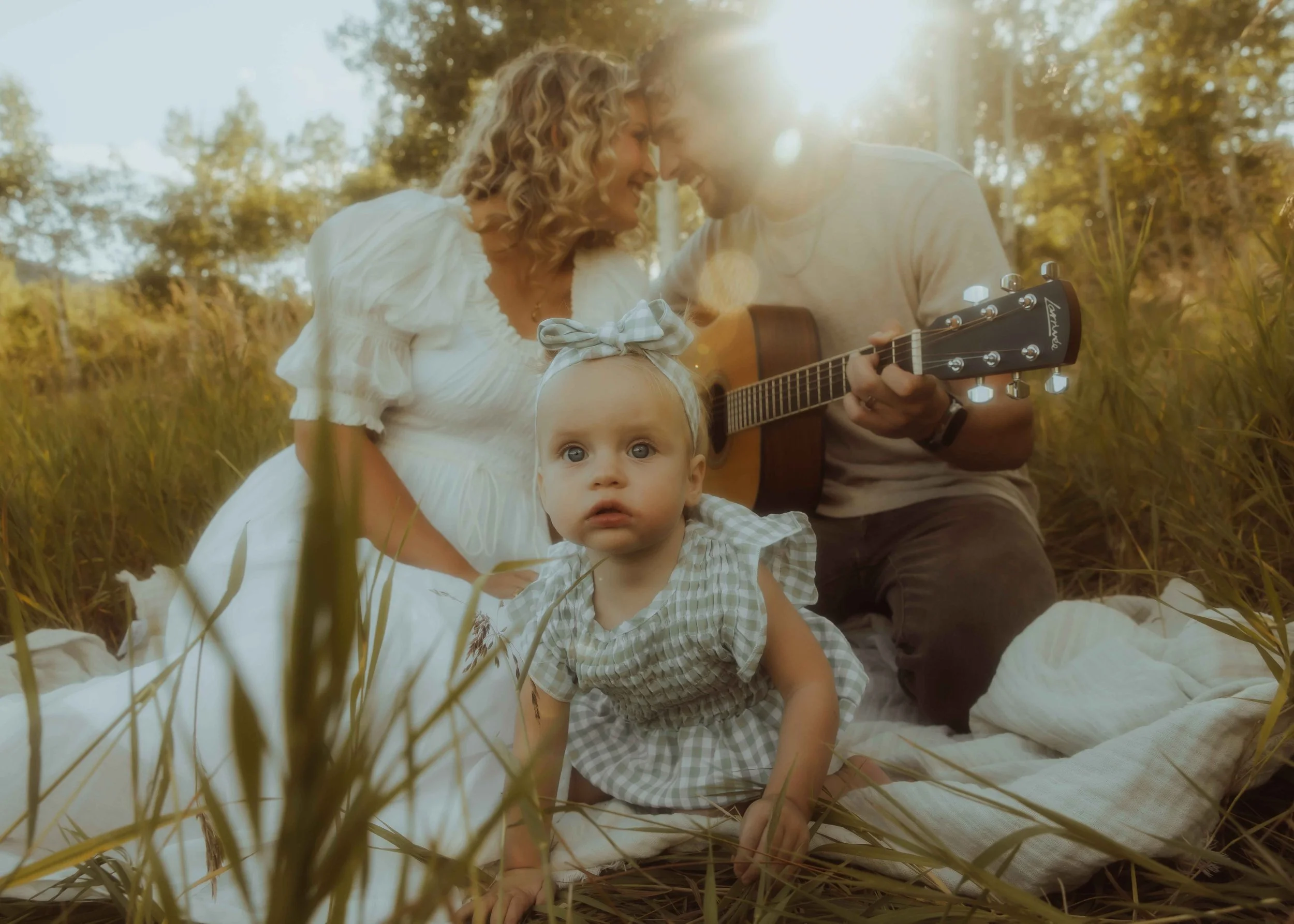 A family of three sitting down on a blanket outside while the father plays the guitar and the baby girl is crawling away