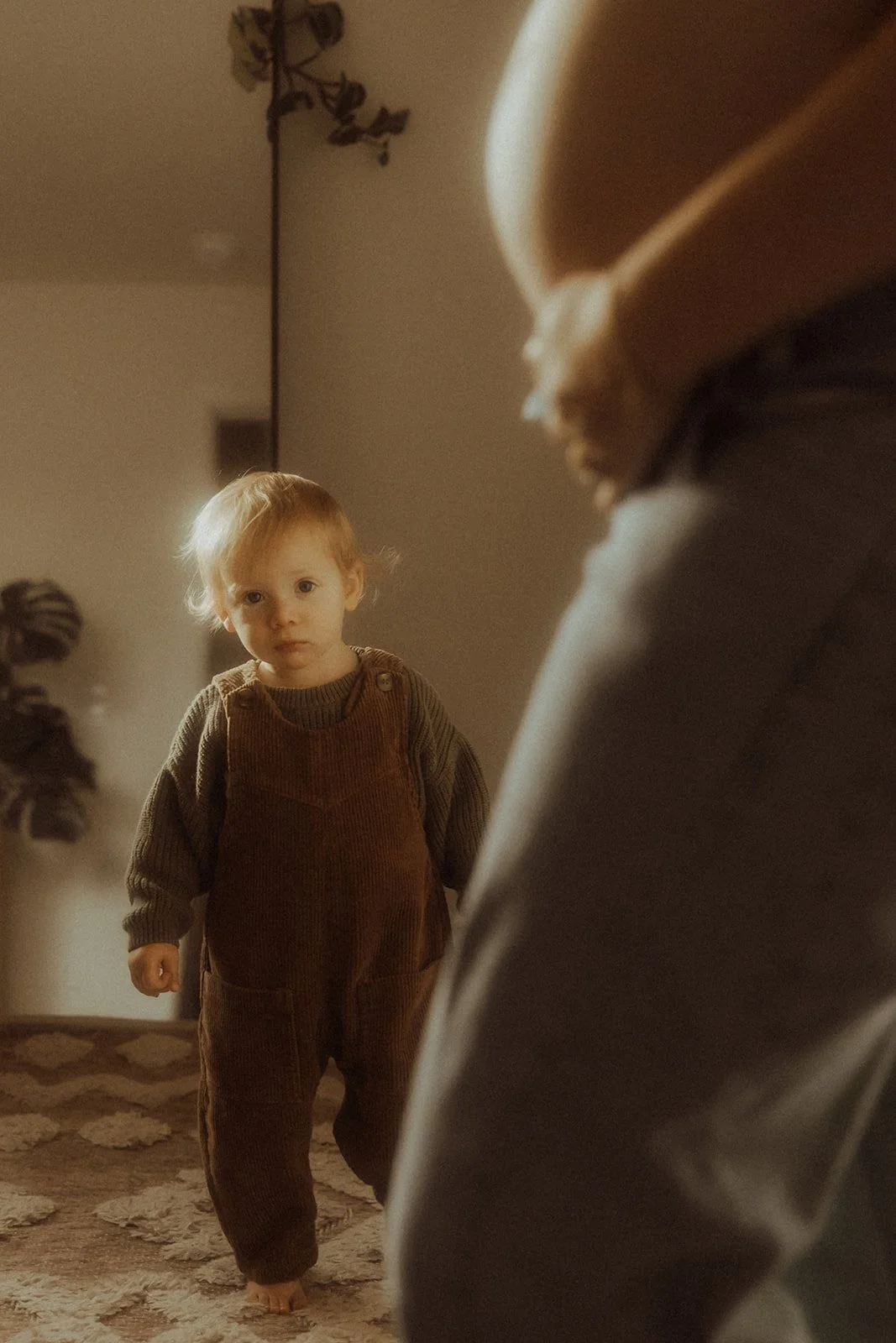 A young boy peaking around his mom's pregnant belly. The soft light from the window creates a warm, cinematic glow in this in-home Utah maternity photo session.