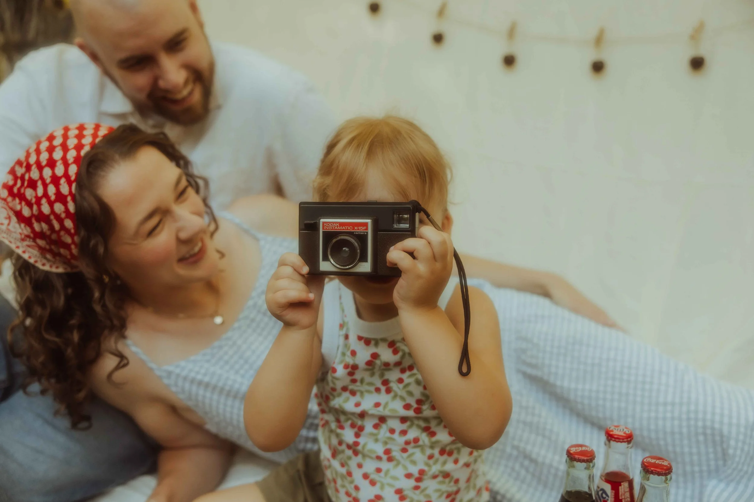 A family of three sitting outside on a linen blanket and the little girl is holding up a vintage camera and smiling.