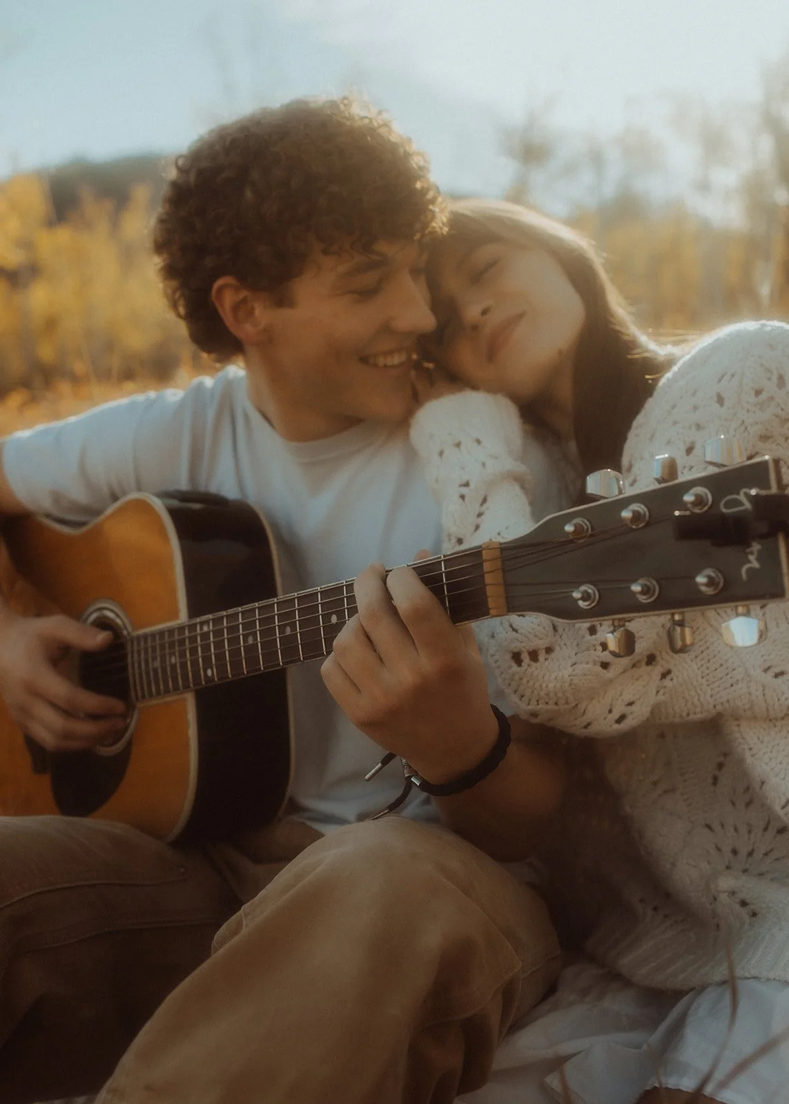 A man and a woman cuddling together while playing the guitar, surrounded by rich fall leaves and colors.