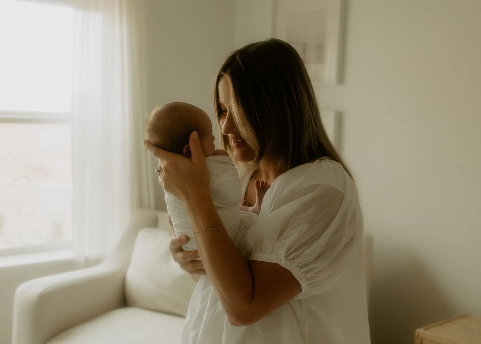 A woman standing in her bedroom holding her newborn baby and smiling down at her baby.