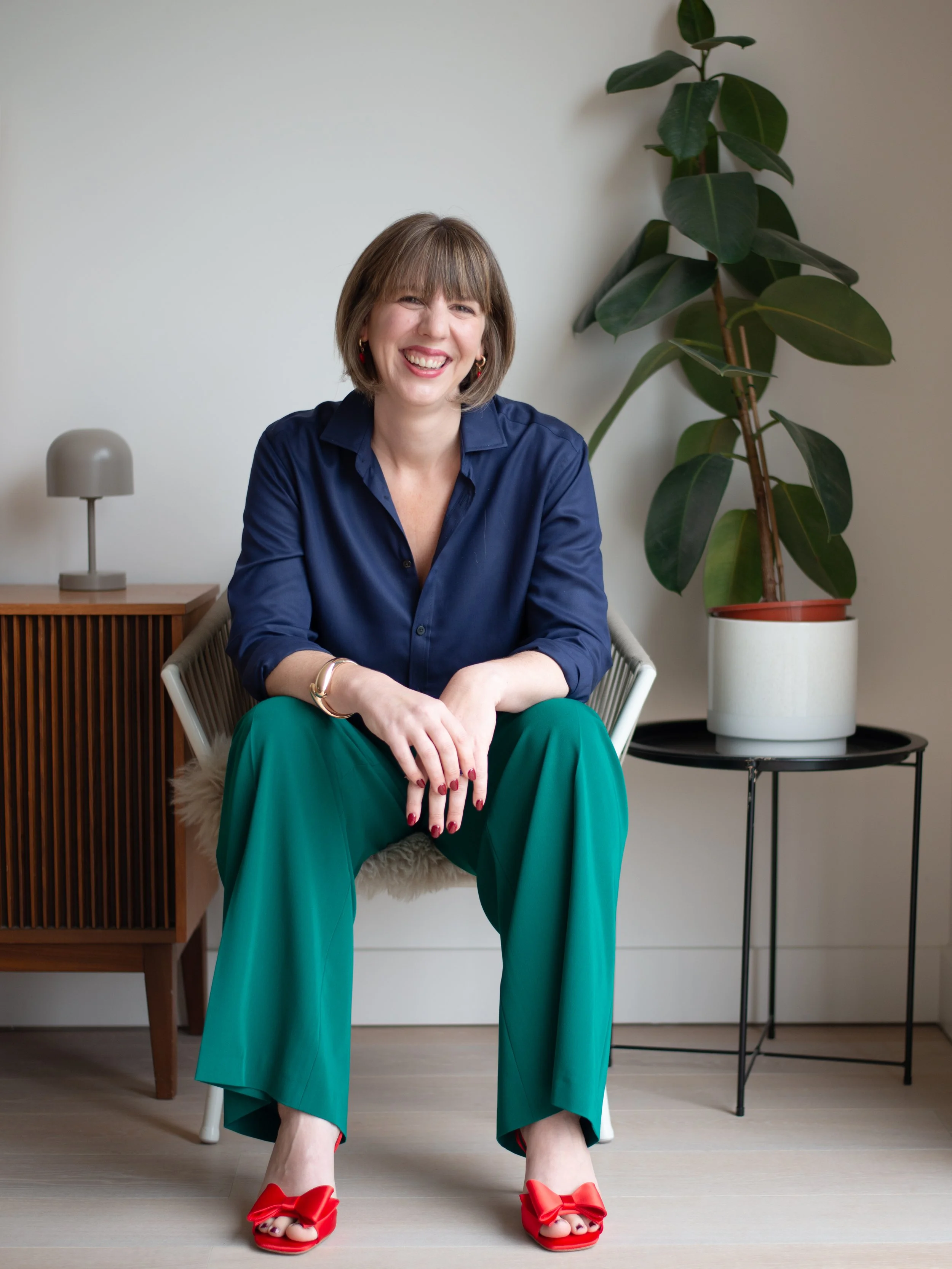 A woman with short brown hair smiling while sitting on a white chair, wearing a navy blue blouse, green wide-leg pants, and red high heel shoes with bows. She is in a room with a wooden side table, a small lamp, and a tall potted plant.