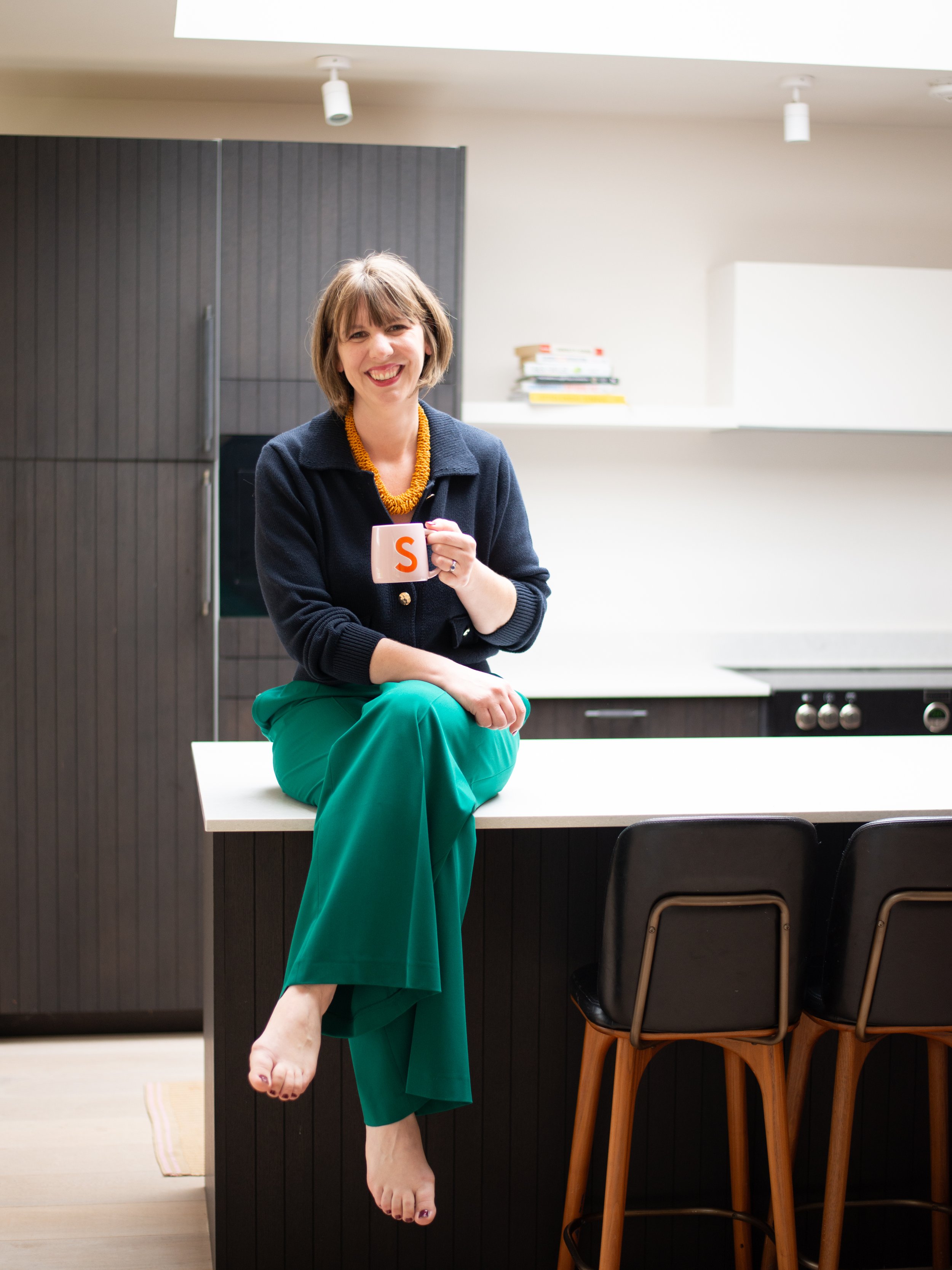 A woman with short brown hair sitting on a kitchen island, smiling, holding a white mug with the letter 'S' on it, in a modern kitchen with black cabinets and chairs.