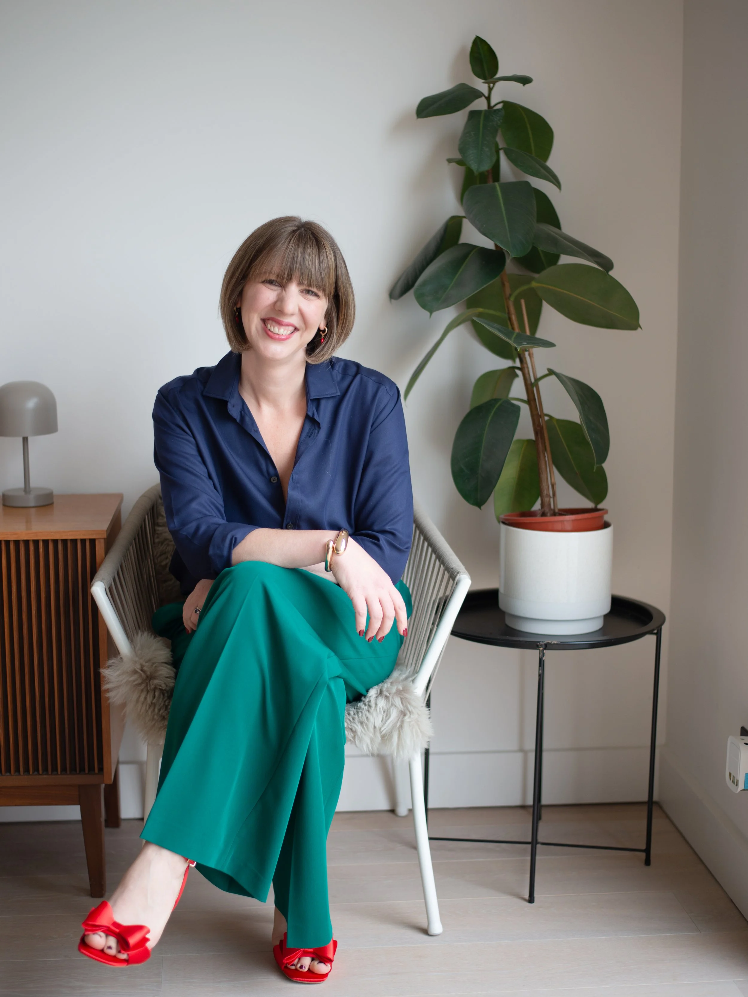 A woman sitting on a white chair with a furry seat cover, smiling, wearing a navy blue blouse, green wide-leg pants, and red high heels with bows. She has short brown hair, hoop earrings, and a bracelet, and is sitting with one leg crossed over the other.