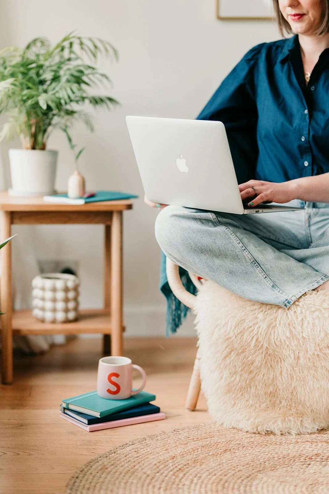A woman sitting on a cozy, plush stool with a sheepskin covering, working on a silver MacBook laptop. She is in a bright, modern room with a wooden floor, a coffee mug, and a small stack of books in front of her. Behind her, there's a small wooden side table with a potted plant, a decorative object, and a closed colorful book.