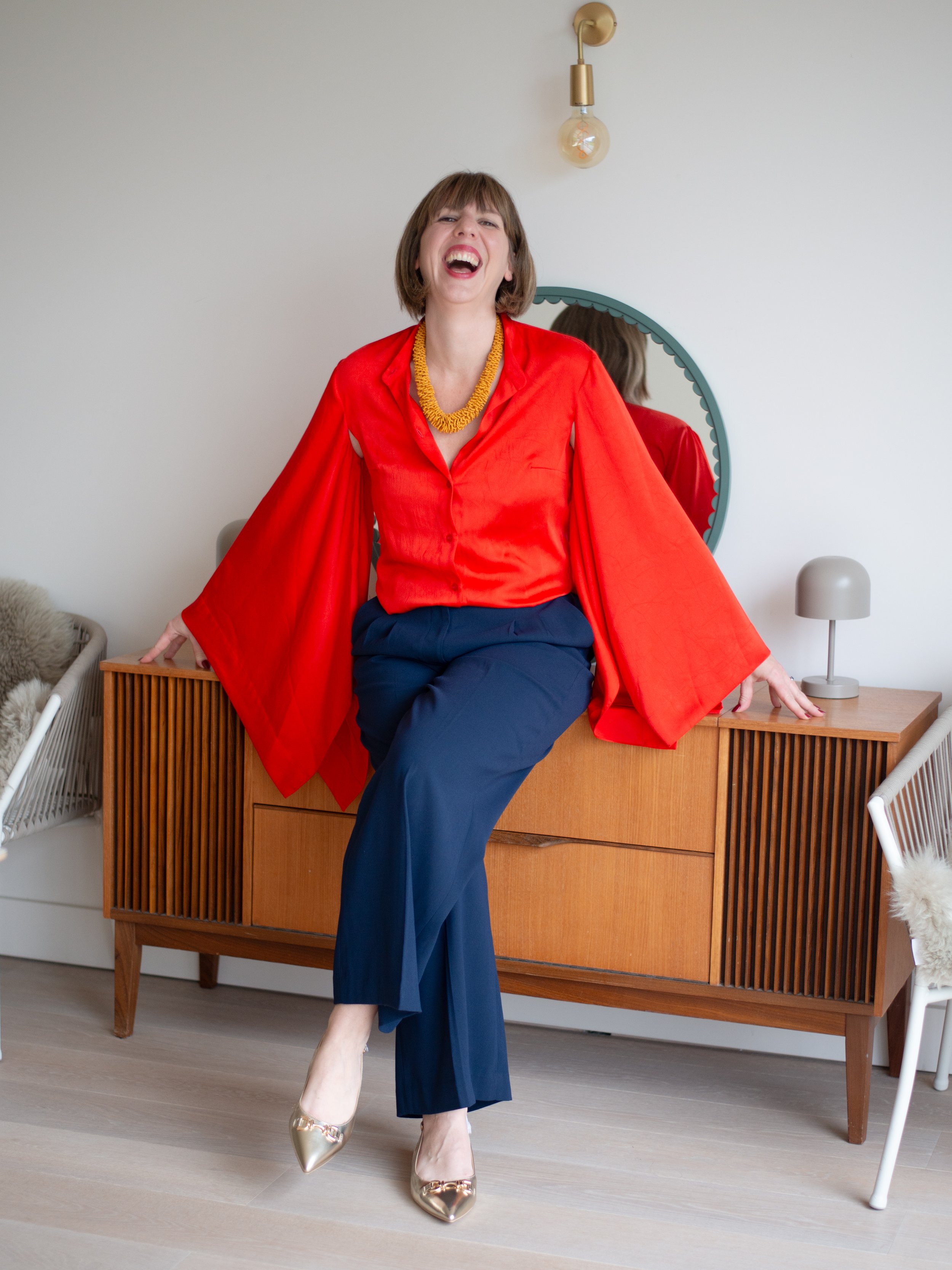 A woman laughing in a stylish red blouse, navy pants, and gold heels, sitting on a wooden sideboard in a modern, minimalistic room with a mirror, lamp, and chairs.