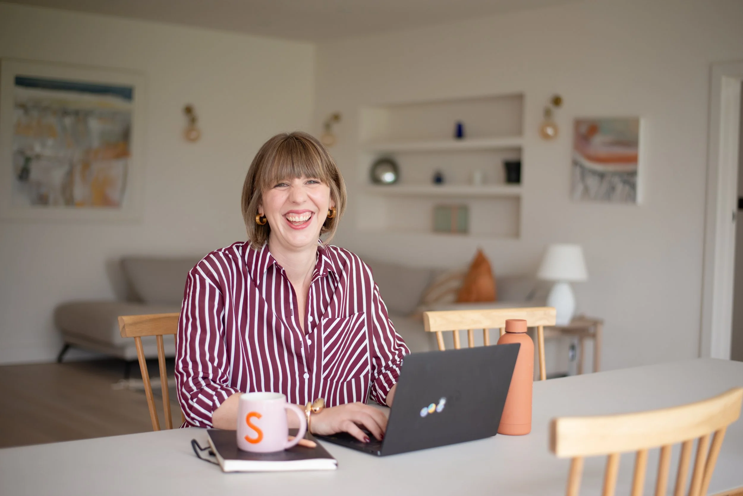A woman with short blonde hair and a striped burgundy and white shirt smiling at a laptop on a dining table in a bright living room.