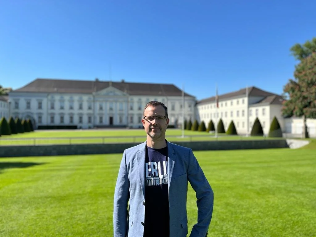 Man standing in front of a large historic building with a manicured lawn, wearing a blue jacket.