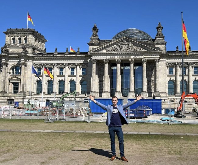 Eine Person, die mit erhobenen Armen vor dem Berliner Reichstagsgebäude steht, mit Baumaschinen im Vordergrund und deutschen Fahnen auf dem Gebäude.
