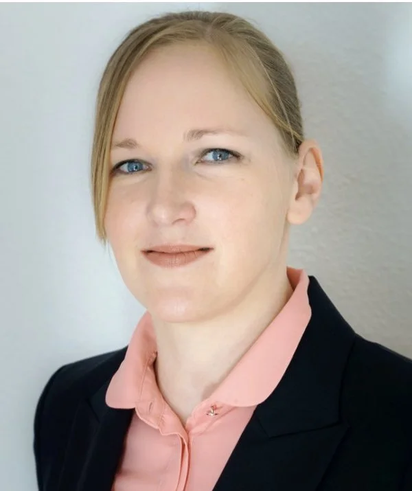 Portrait of a woman in a black blazer and peach blouse, with light-colored hair styled to the side, against a plain background.