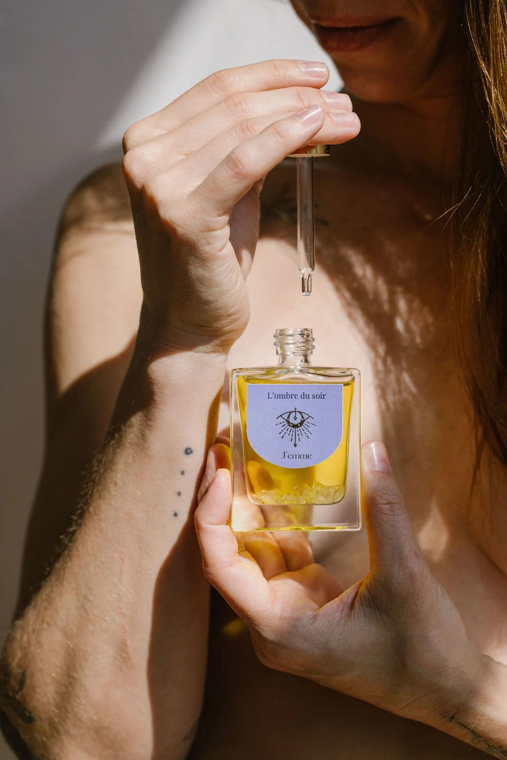 Une femme tient un flacon de parfum jaune, intitulé "L'ombre du soir" avec une étiquette mentionnant "J'aime". Elle verse le parfum à l'aide d'une pipette.