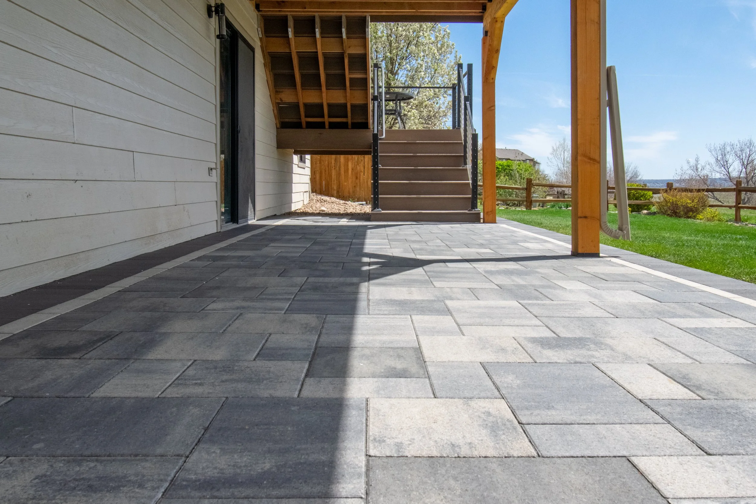 View of a backyard patio with gray pavers, a sliding glass door, and a wooden staircase leading up to a deck. There is a grassy yard with a wooden fence and trees in the background.