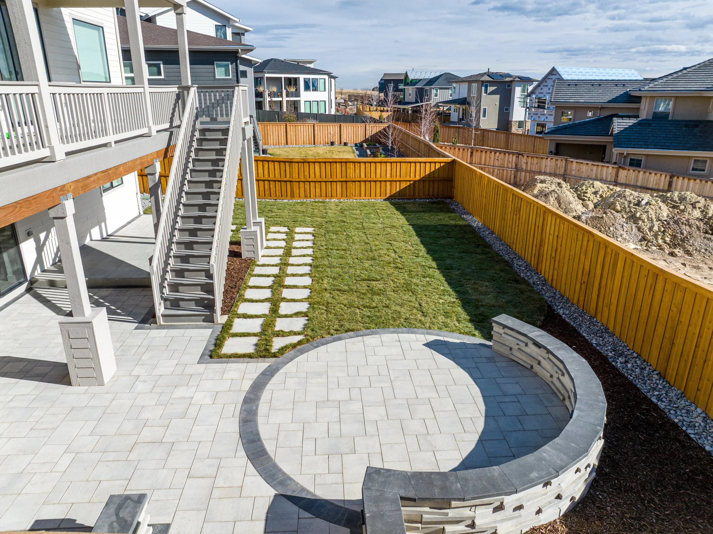 A backyard with a patio with hardscape paver patio, seated bench, deck and landscape lighting