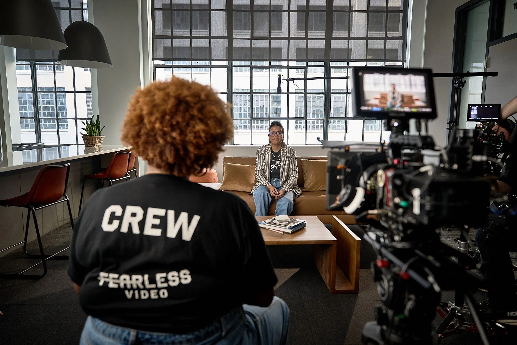A woman with glasses and a striped jacket sitting on a couch in a modern room with large windows while being filmed. A crew member with curly hair and a black shirt with 'CREW' printed on the back is in the foreground, and professional video equipment is visible on the right.
