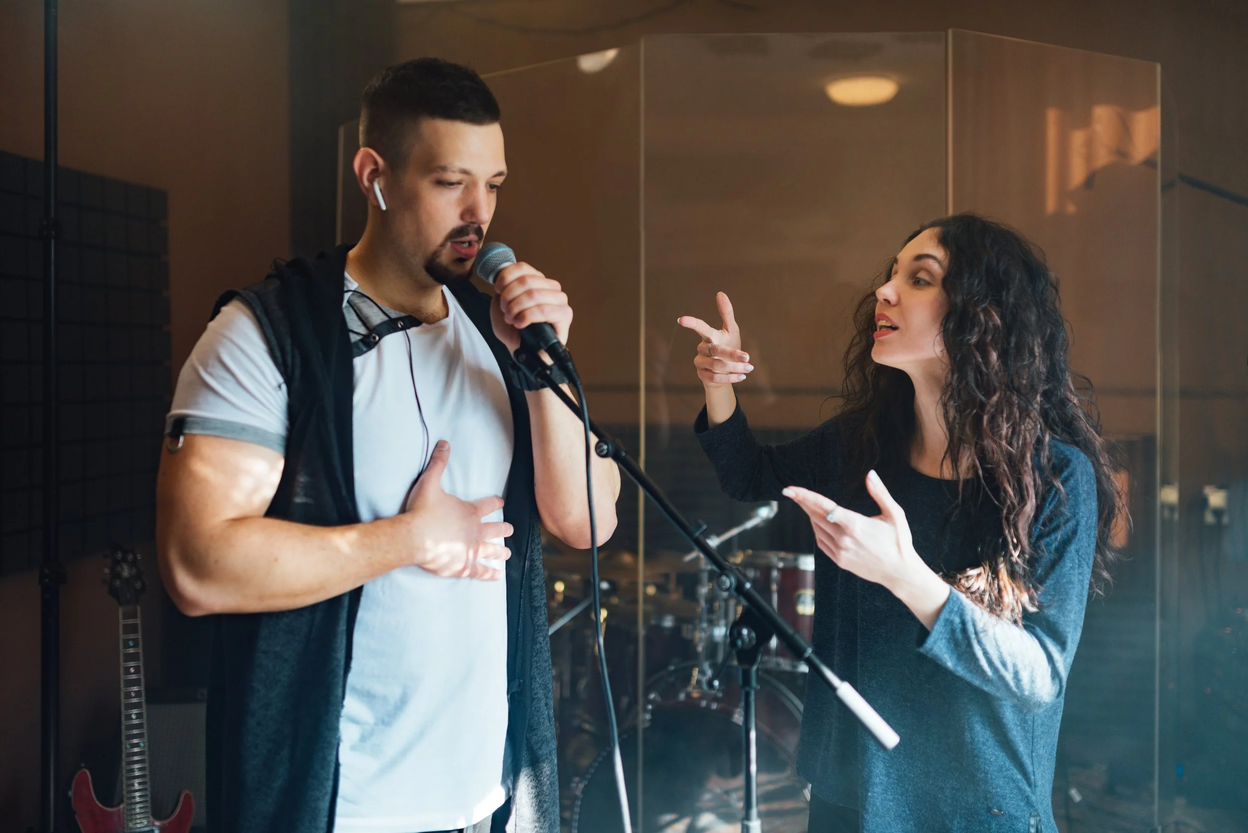 A man sings into a microphone with his hand on his chest, while a woman gestures animatedly and talks to him during a music rehearsal or recording session in a studio with musical instruments behind them.