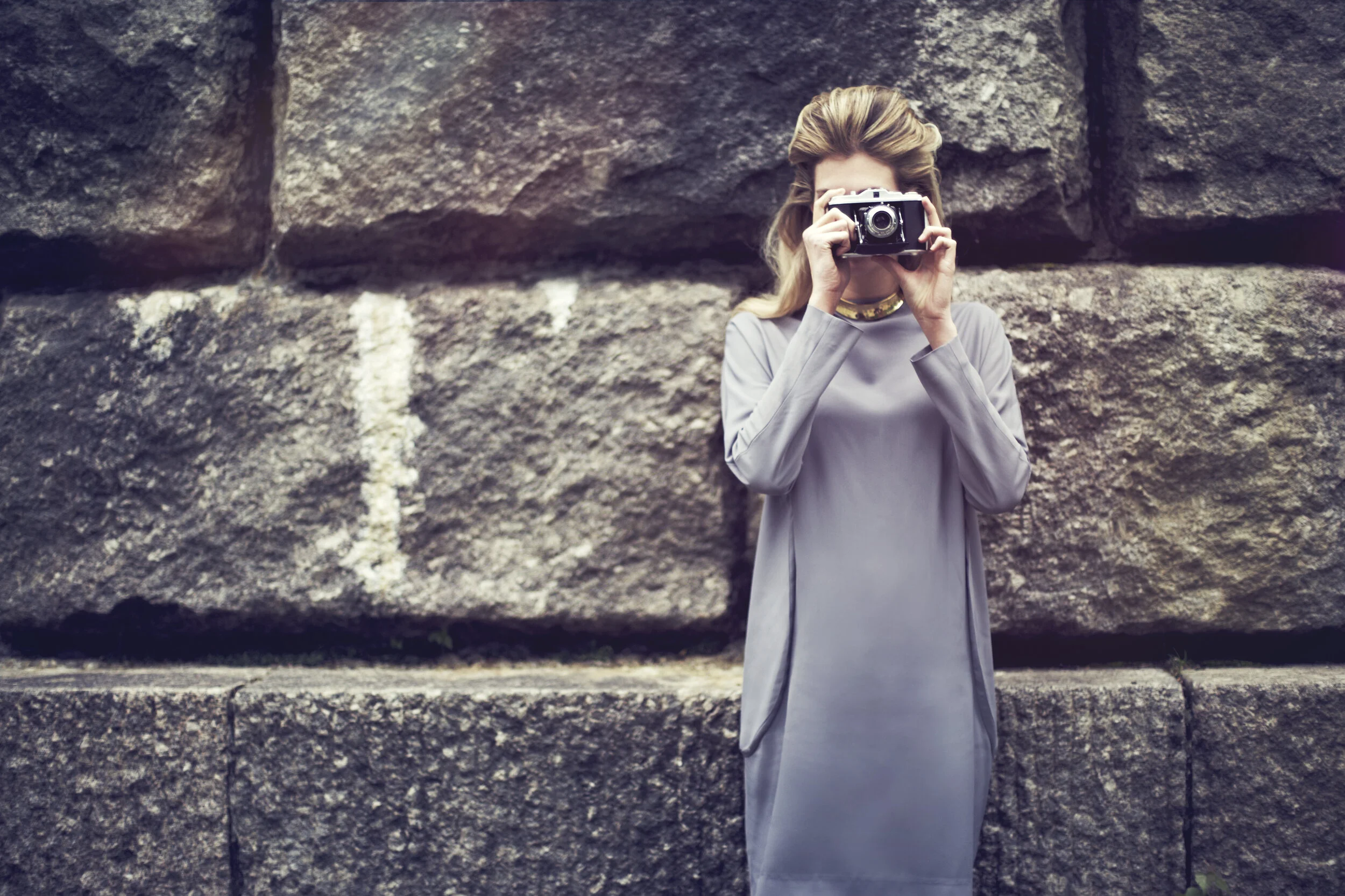 Ein Modefoto zeigt eine Frau in einem hochwertigem Outfit, das Gesicht wird von einer Kamera verdeckt. Im Hintergrund ist eine Monumentale Mauer