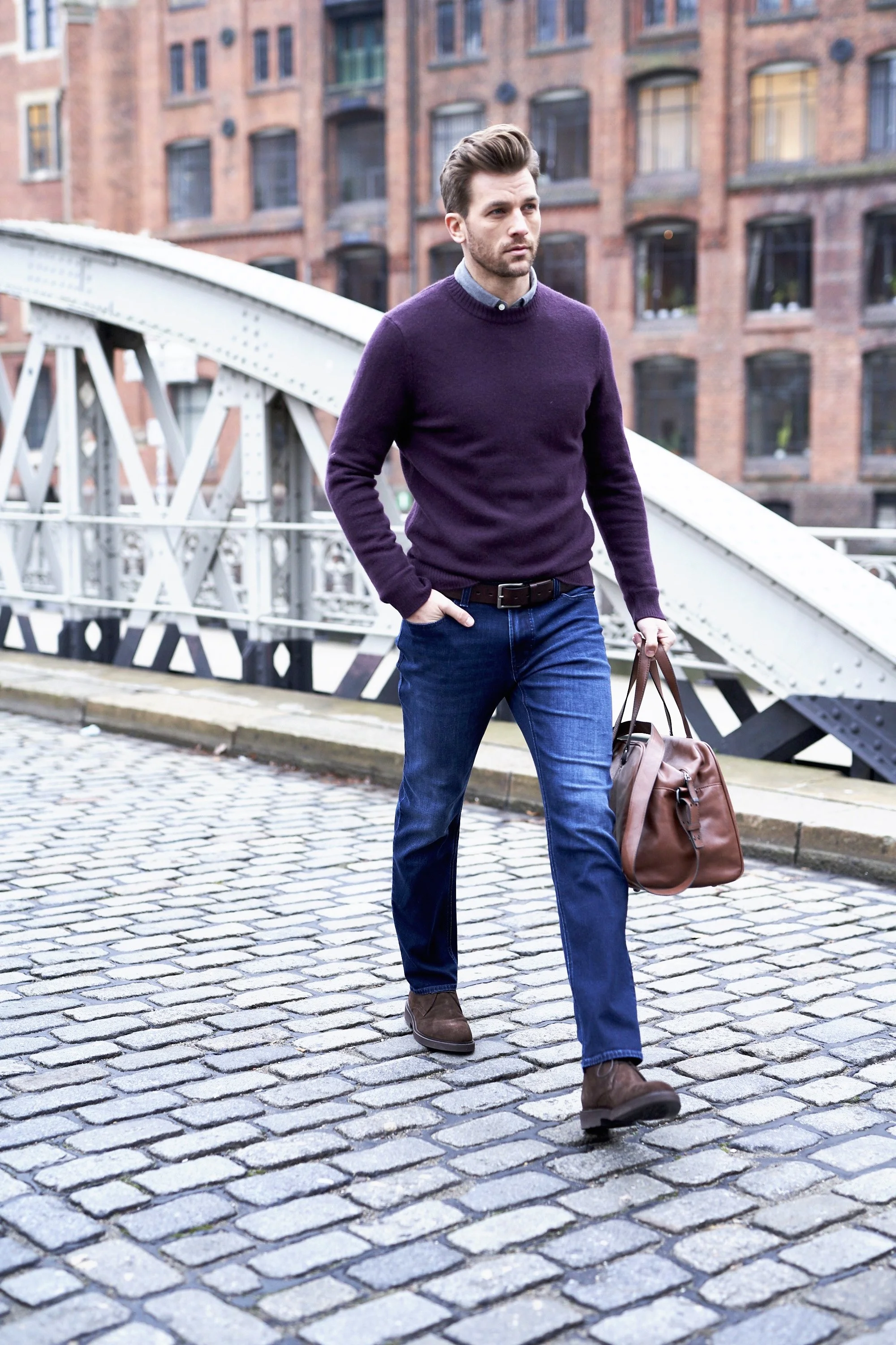 Ein Modefoto. Mann überquert eine Straße mit Kopfsteinpflaster und trägt einen Weekender aus Leder bei sich