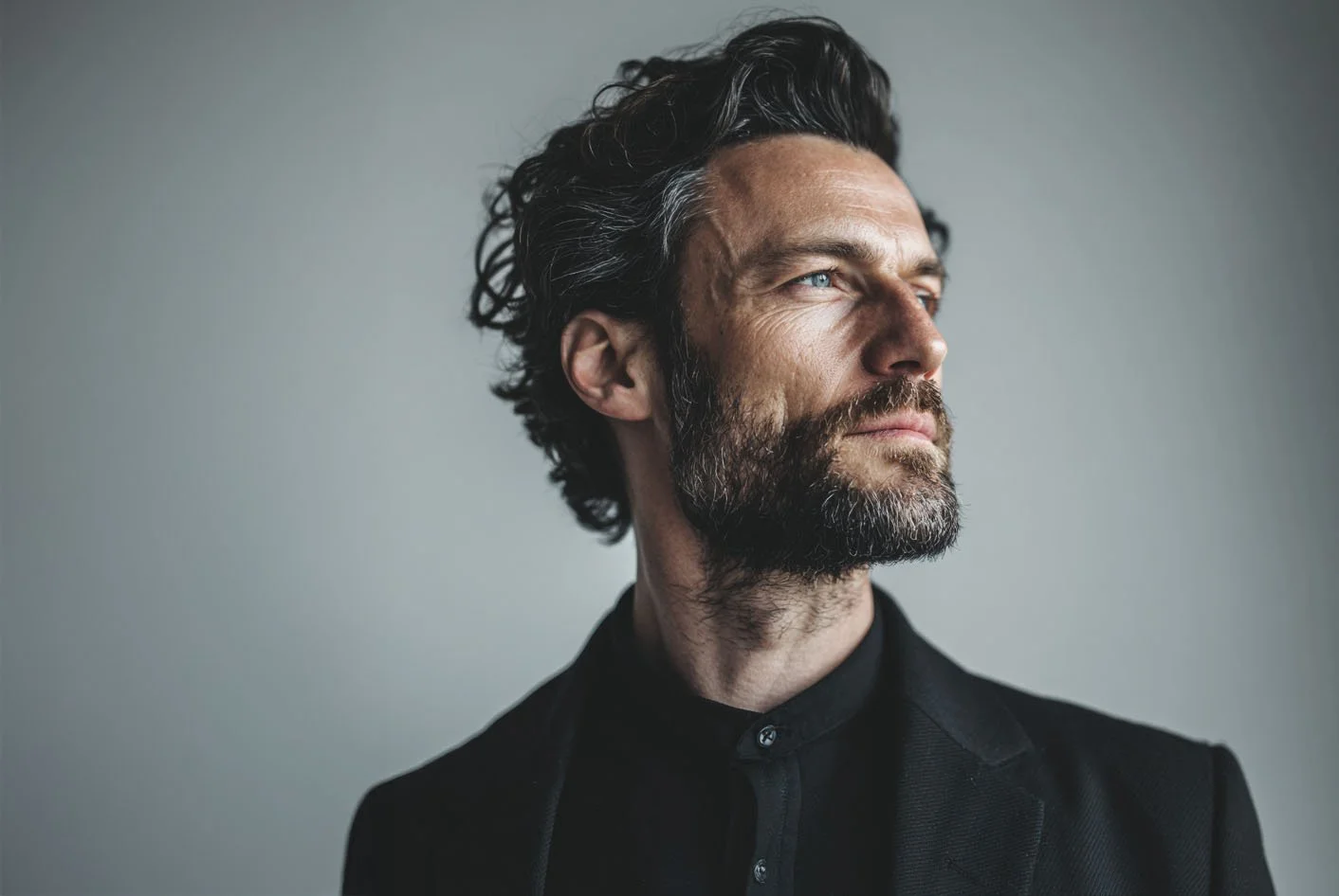 Nahaufnahme eines Mannes mit dunklem, grauem Haar und Bart, blickt nach rechts, trägt einen schwarzen Anzug, neutraler Hintergrund.