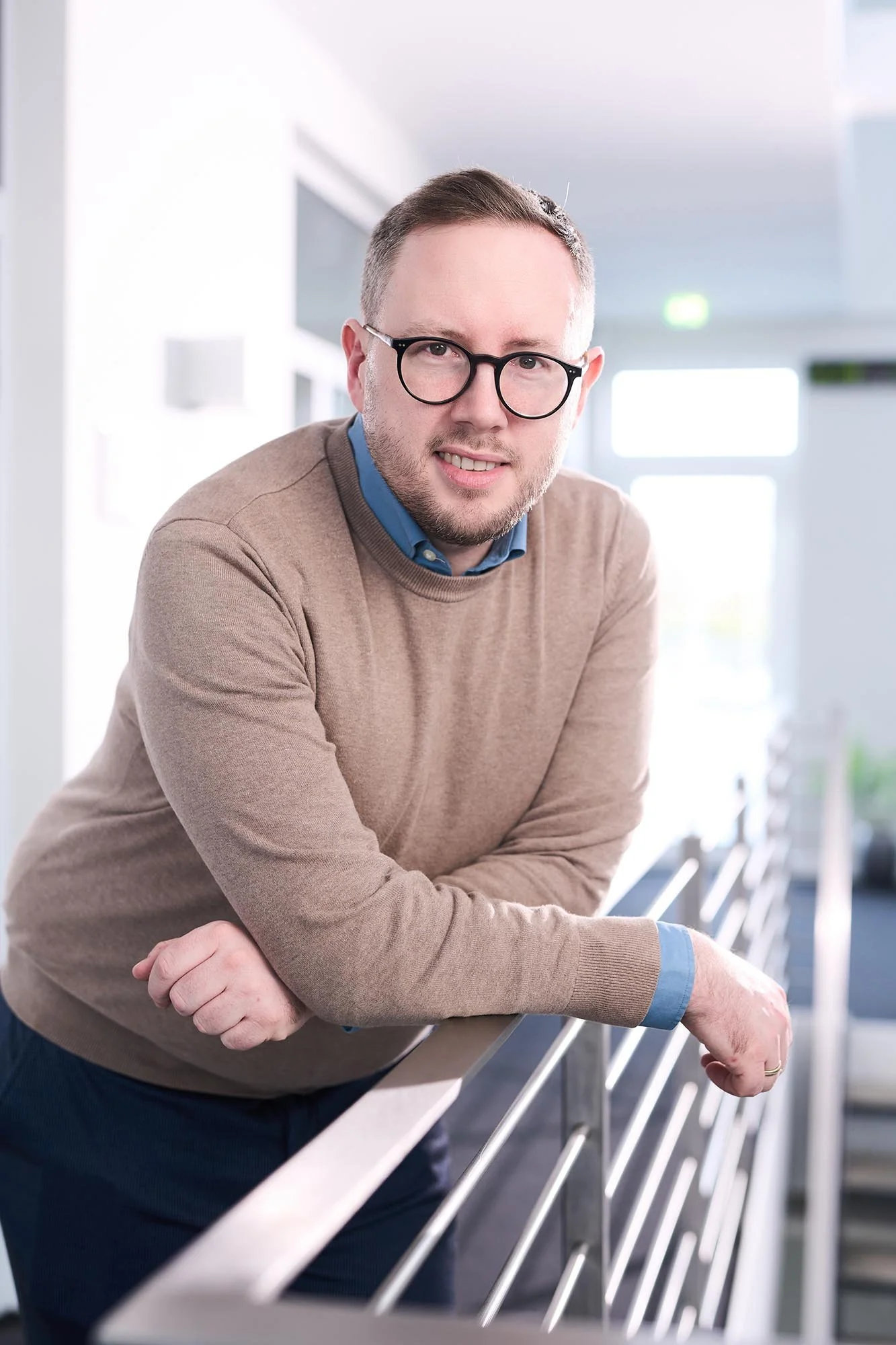 Ein Business Foto eines Mannes mit Brille lehnt an einem Geländer in einem modernen Innenraum. 
