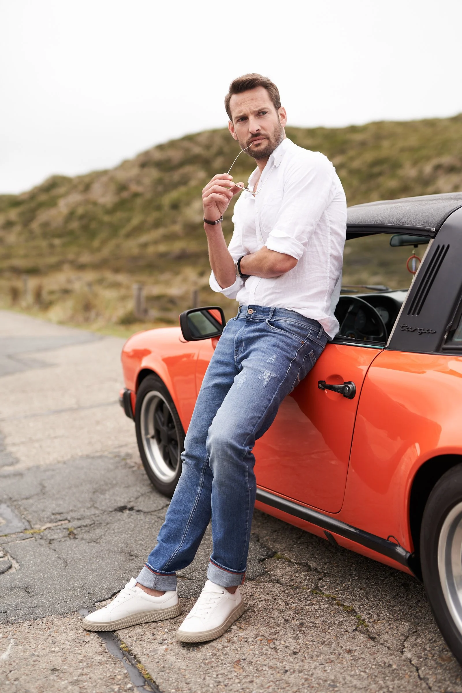 Ein Modefoto eines Mannes der an einem orangenem Porsche 911 angelehnt steht