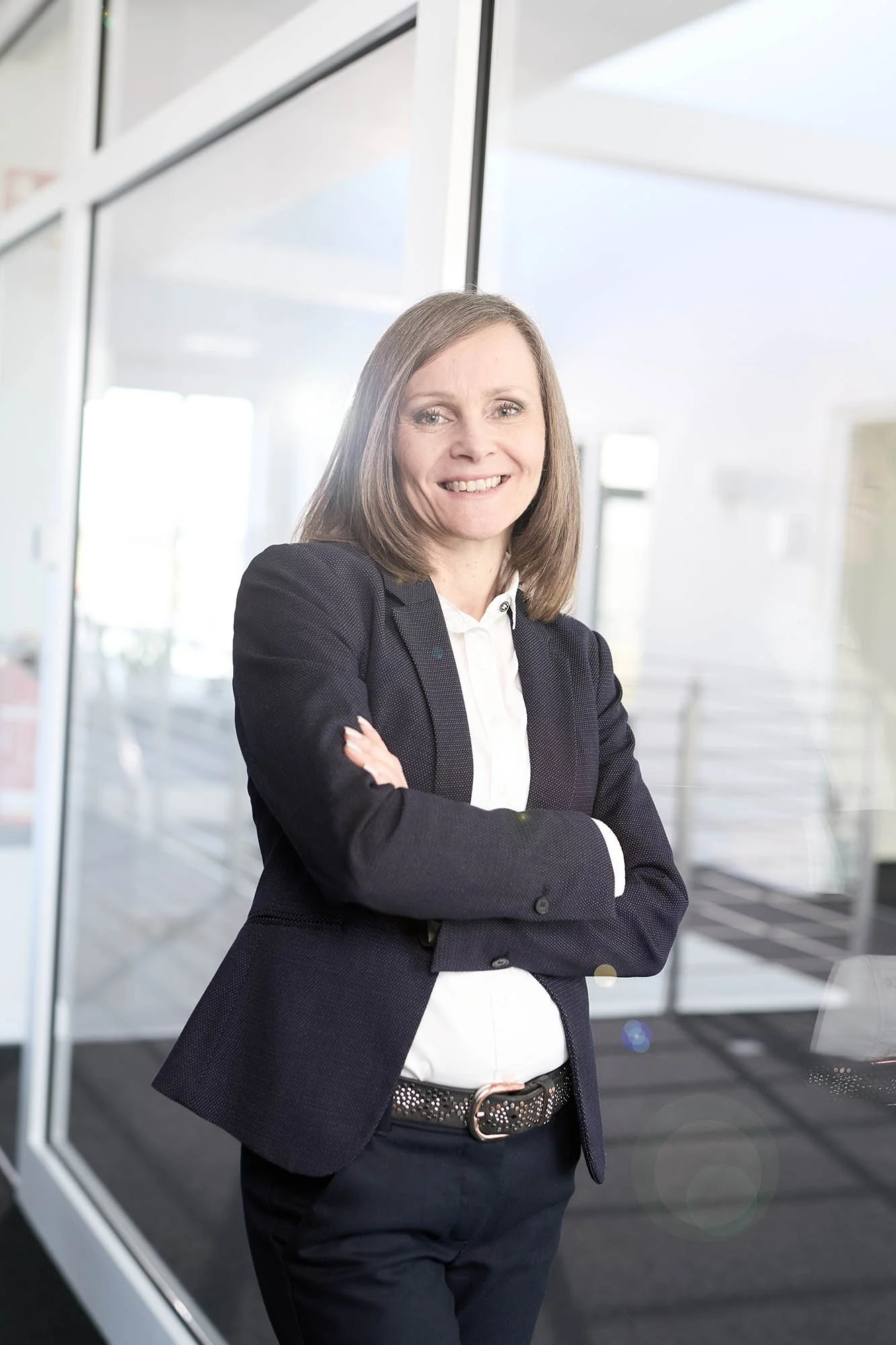 Eine lächelnde Frau in formeller Kleidung steht mit verschränkten Armen in einem modernen Bürogebäude.