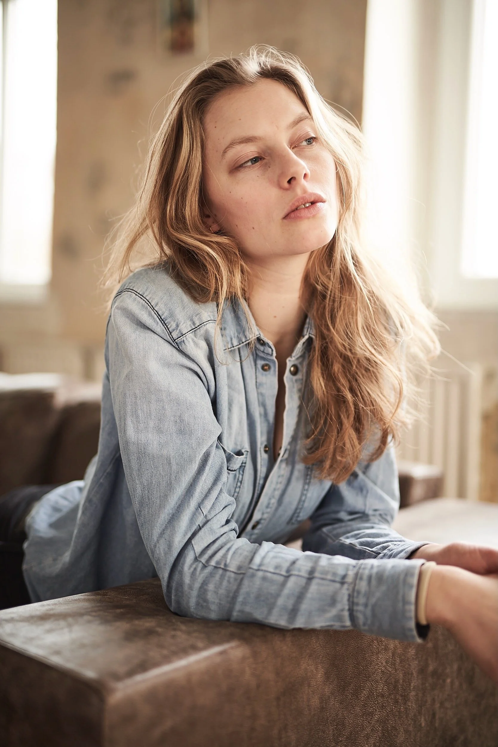 Eine Frau mit langen, blonden Haaren, die ein Jeanshemd trägt und auf einem Sofa lehnt. Im Hintergrund ist ein unscharfer Raum zu sehen.