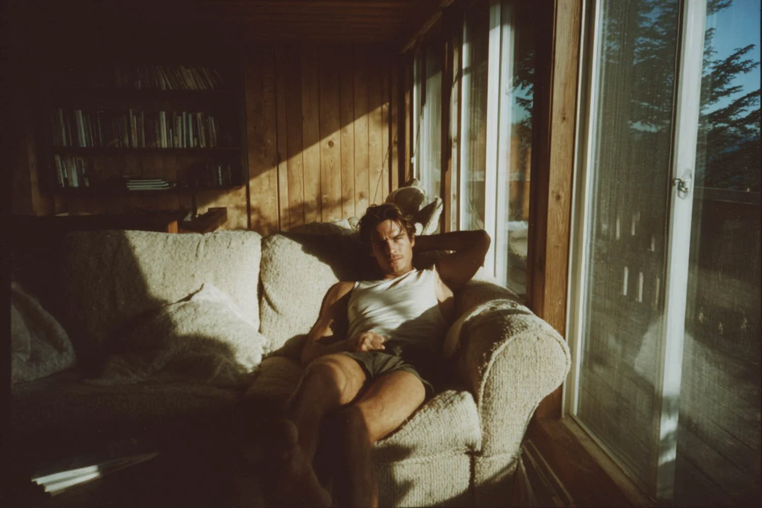 Junger Mann liegt entspannt auf einem gemütlichen Sofa in einem Raum mit Holzwandpaneelen, Sonnenlicht scheint durch große Fenster und wirft Schatten im Raum.