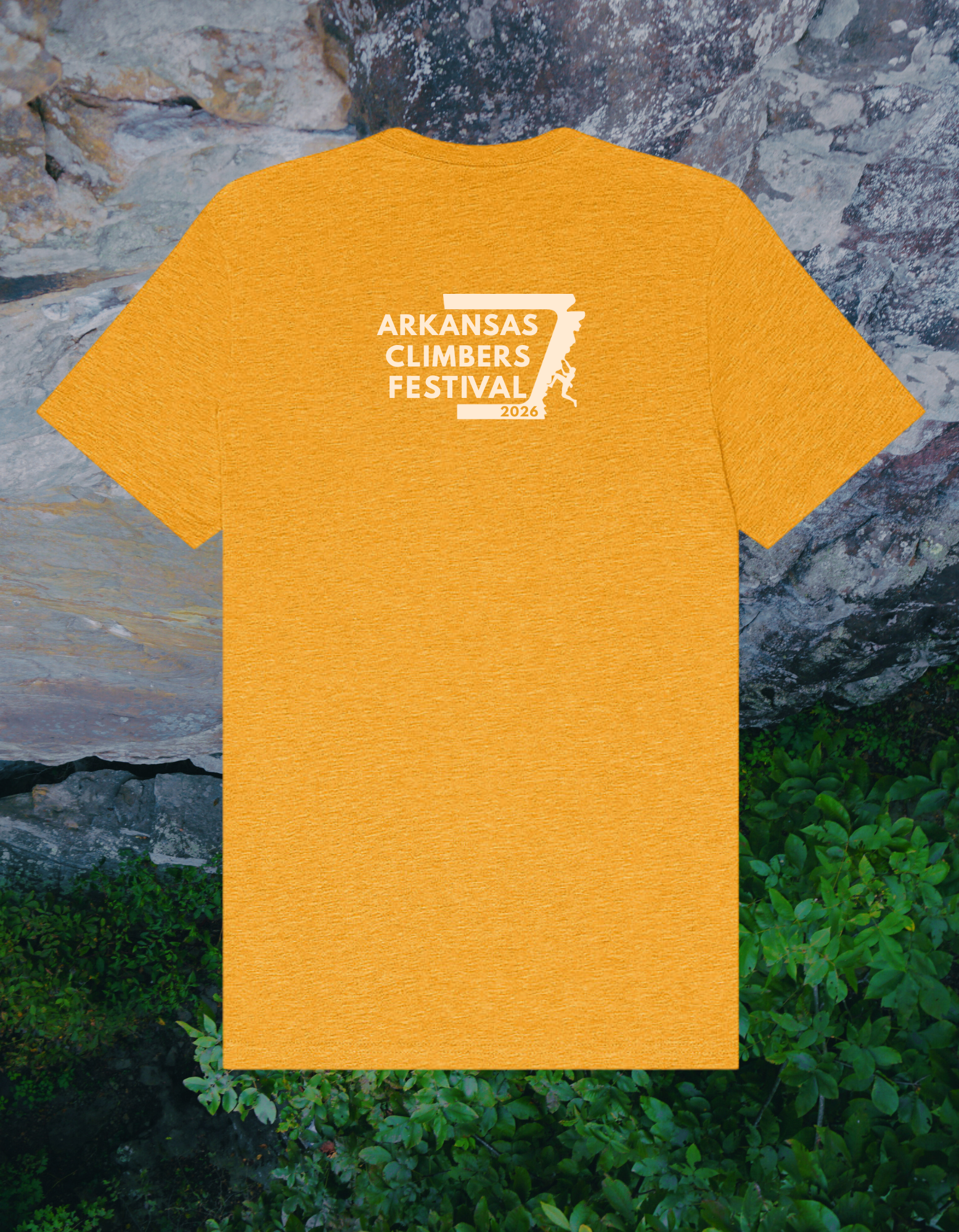 Yellow T-shirt featuring a logo for the Arkansas Climbers Festival 2026, with text and a mountain climber graphic, hanging outdoors against a rocky background with green foliage.