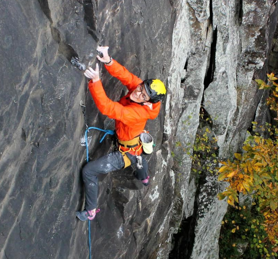 A smiling woman wearing a white climbing helmet with teal accents, black shirt, and a gold chain necklace, standing in front of a rocky cliff with climbing ropes draped over her shoulders.