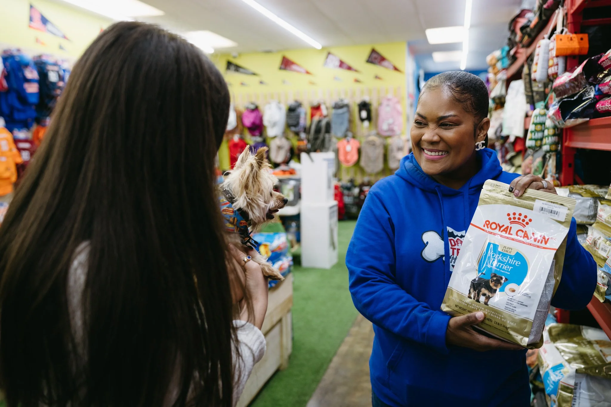 One of the owners of Dawg Team shows some dog food products to a pet owner, who is holding their Yorkshire Terrier. They are chatting and smiling together in the store.