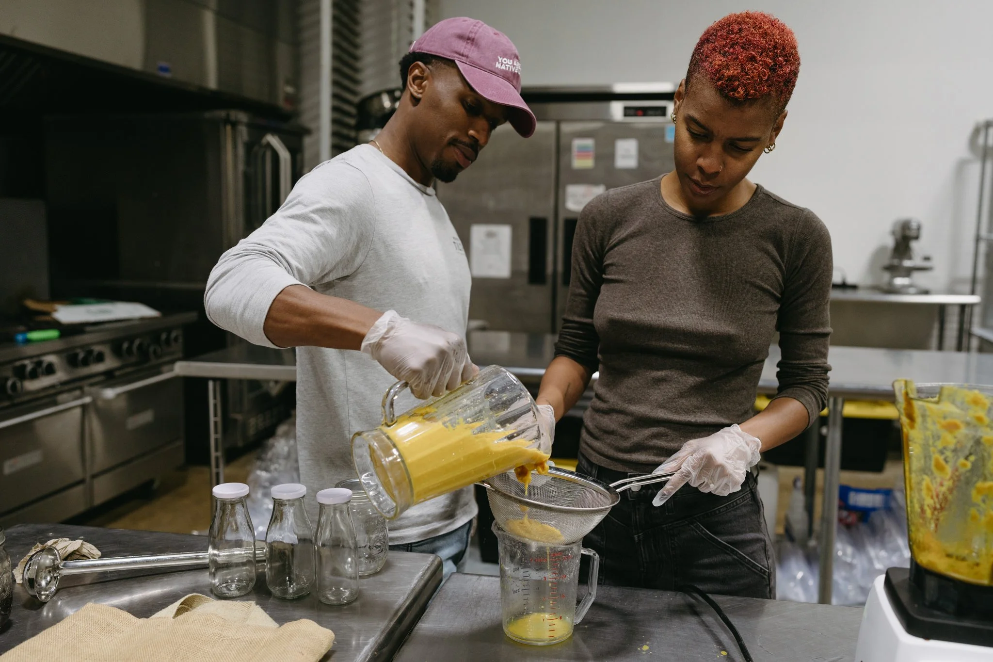 Talibah and Bertrum are making ginger and turmeric juice in their shared commercial kitchen. Bertrum is pouring the ginger pulp through a sieve whilst Talibah holds the jug that the juice is going into.