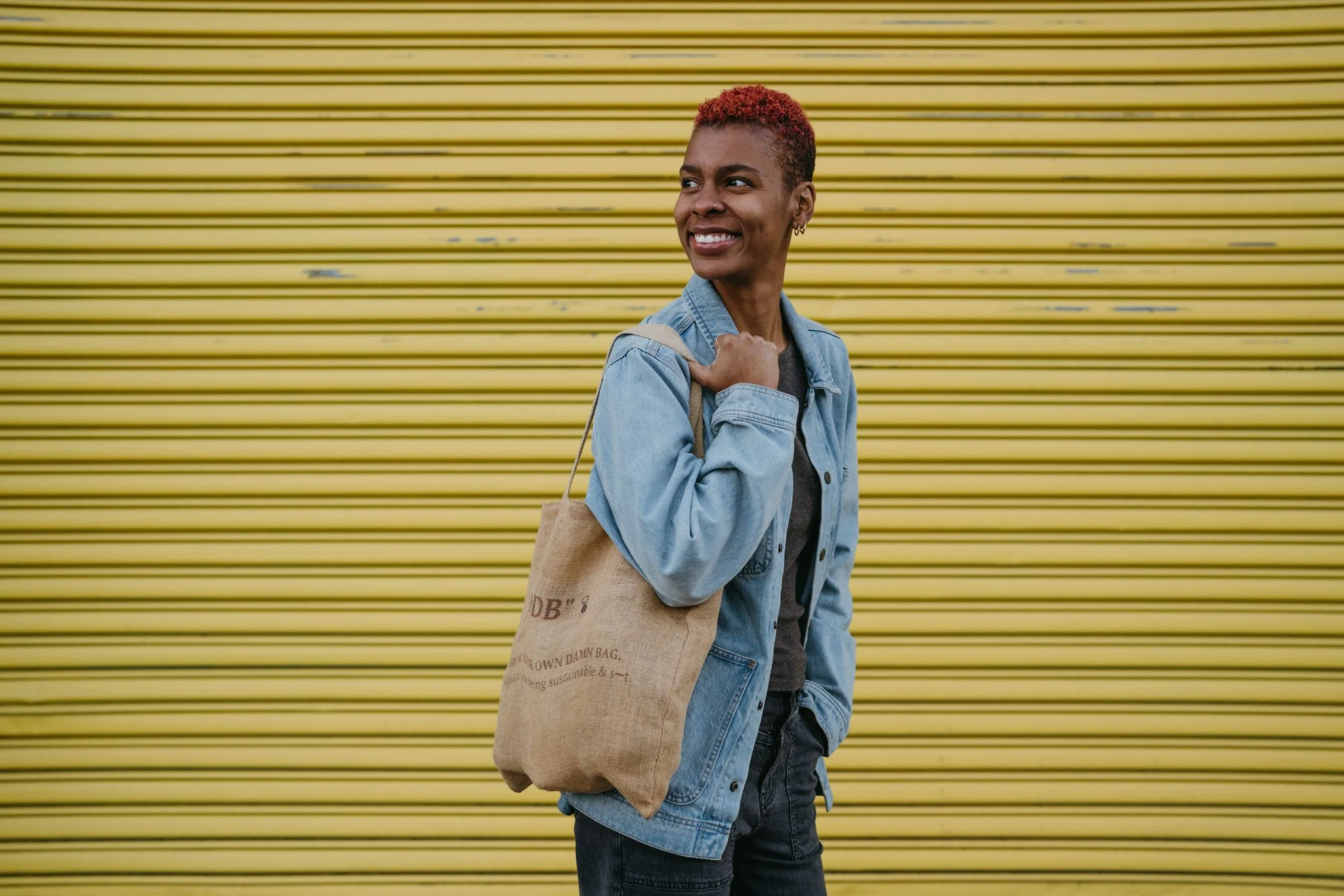 Talibah, the founder of Mama's Sundry, models a reusable bag made by the brand. Talibah is wearing a blue denim jacket and has red hair, the backdrop is a metal, bright yellow shutter.