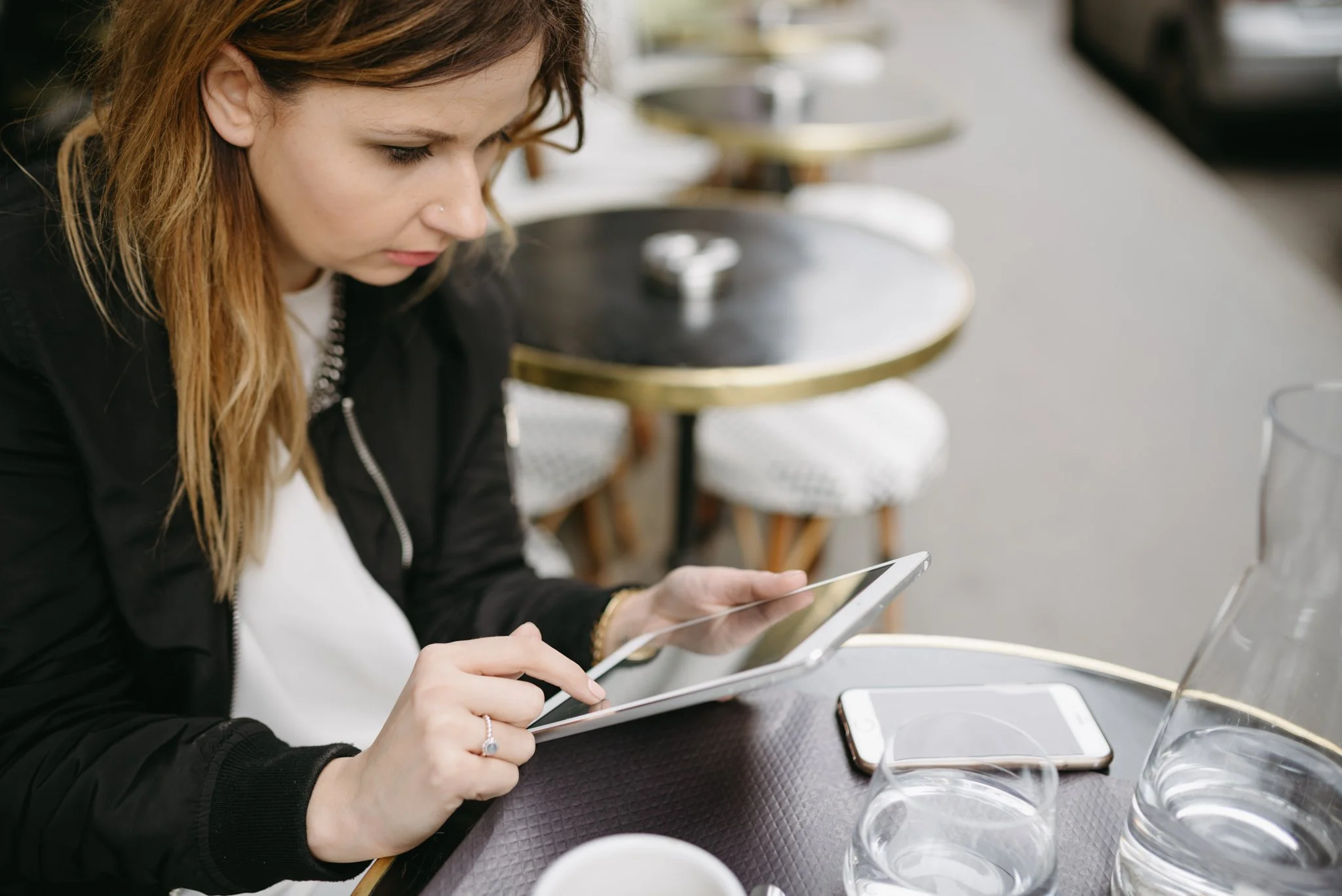 A portrait of Iga, a coach and mentor sat at a café table in the centre of Paris. She is doing work on her iPad whilst drinking a coffee, whilst her phone is on the table.