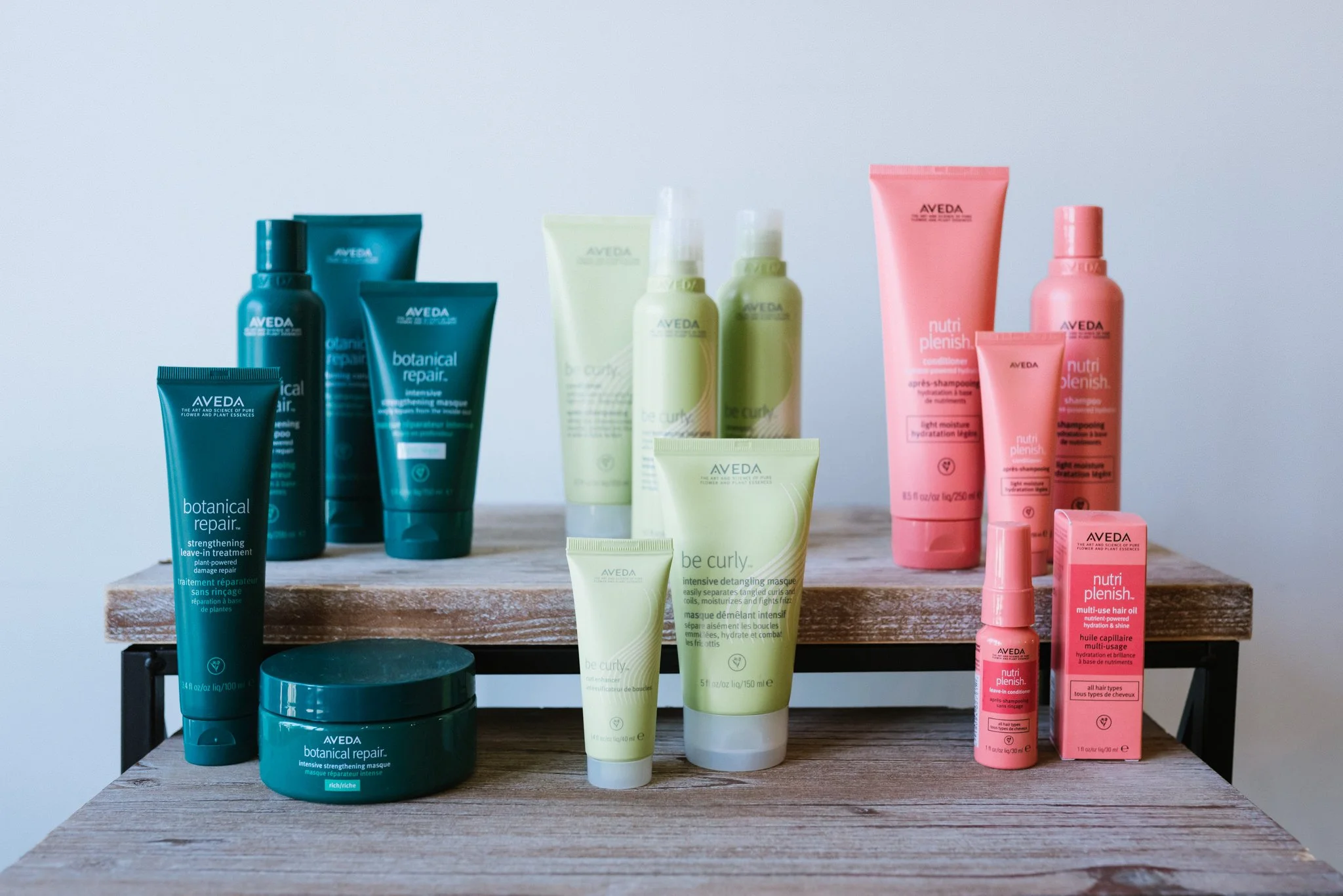 A group of Aveda hair products are photographed on a wooden shelf with a white wall behind them.
