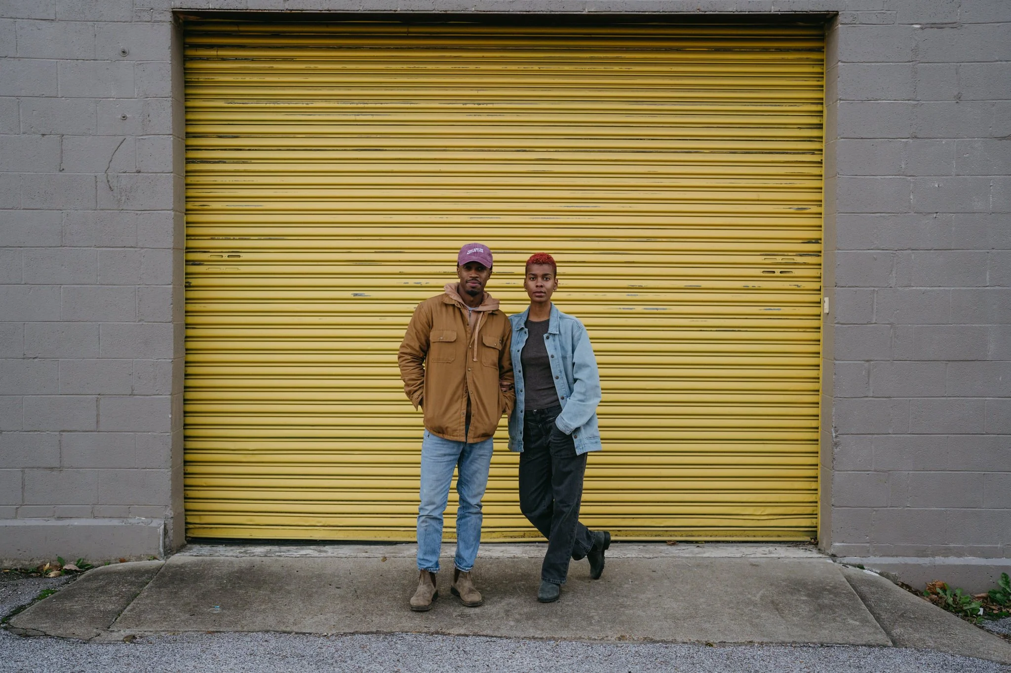 Talibah and Bertrum are posing for a portrait together infront of a large bright yellow metal shutter. 