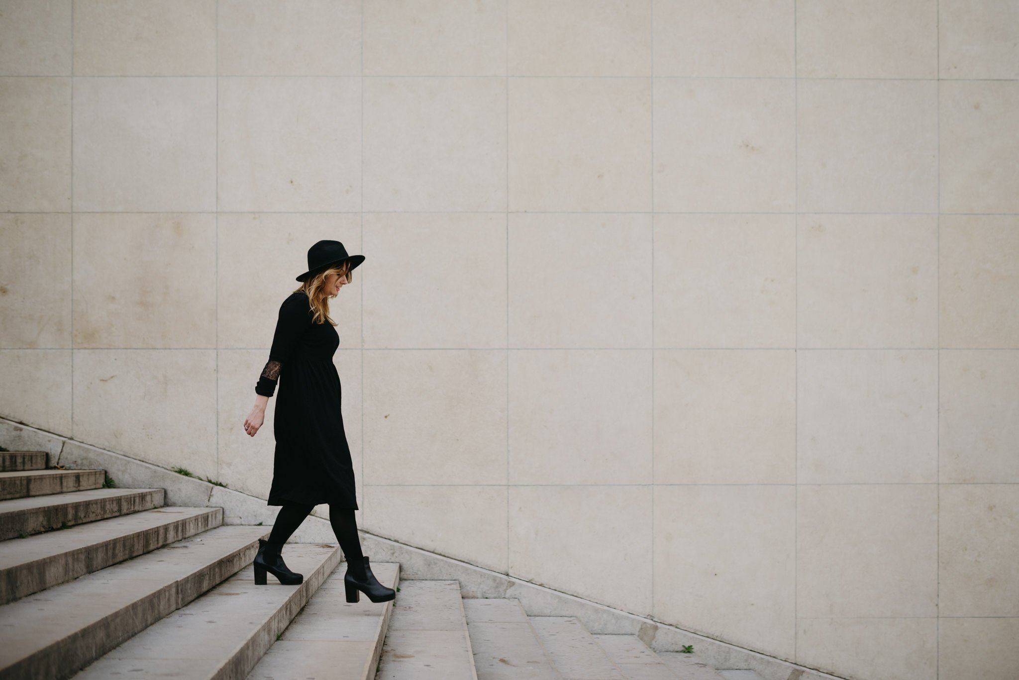 A photo of Iga, a coach and mentor, walking down the stairs at Trocadero in Paris near the Eiffel Tower. She is dressed all in black, with a wide brimmed black hat, and wavy blonde hair.