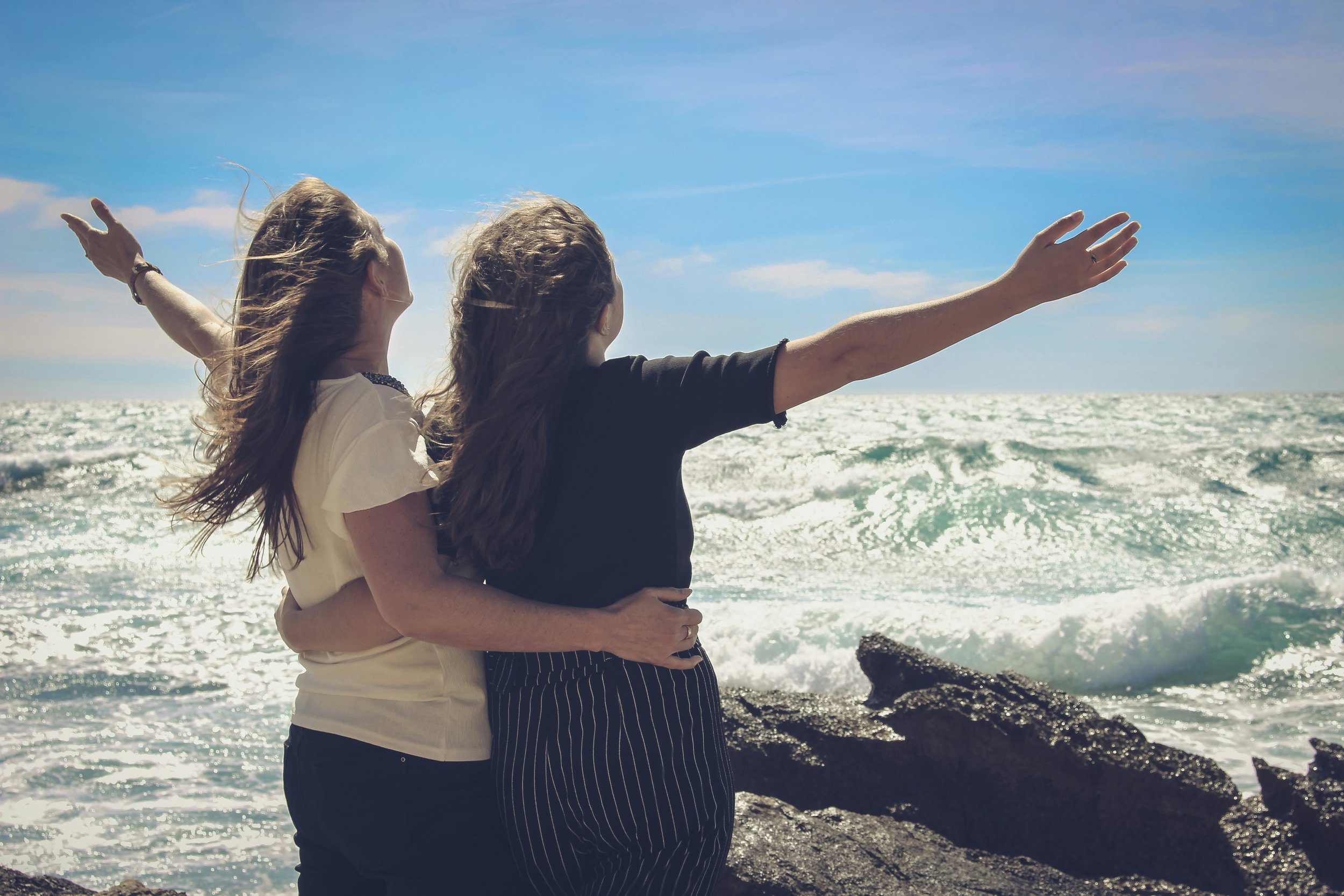 Two women at the beach, facing the ocean, one with arms outstretched, and the other holding her from behind, enjoying a sunny day by the water.