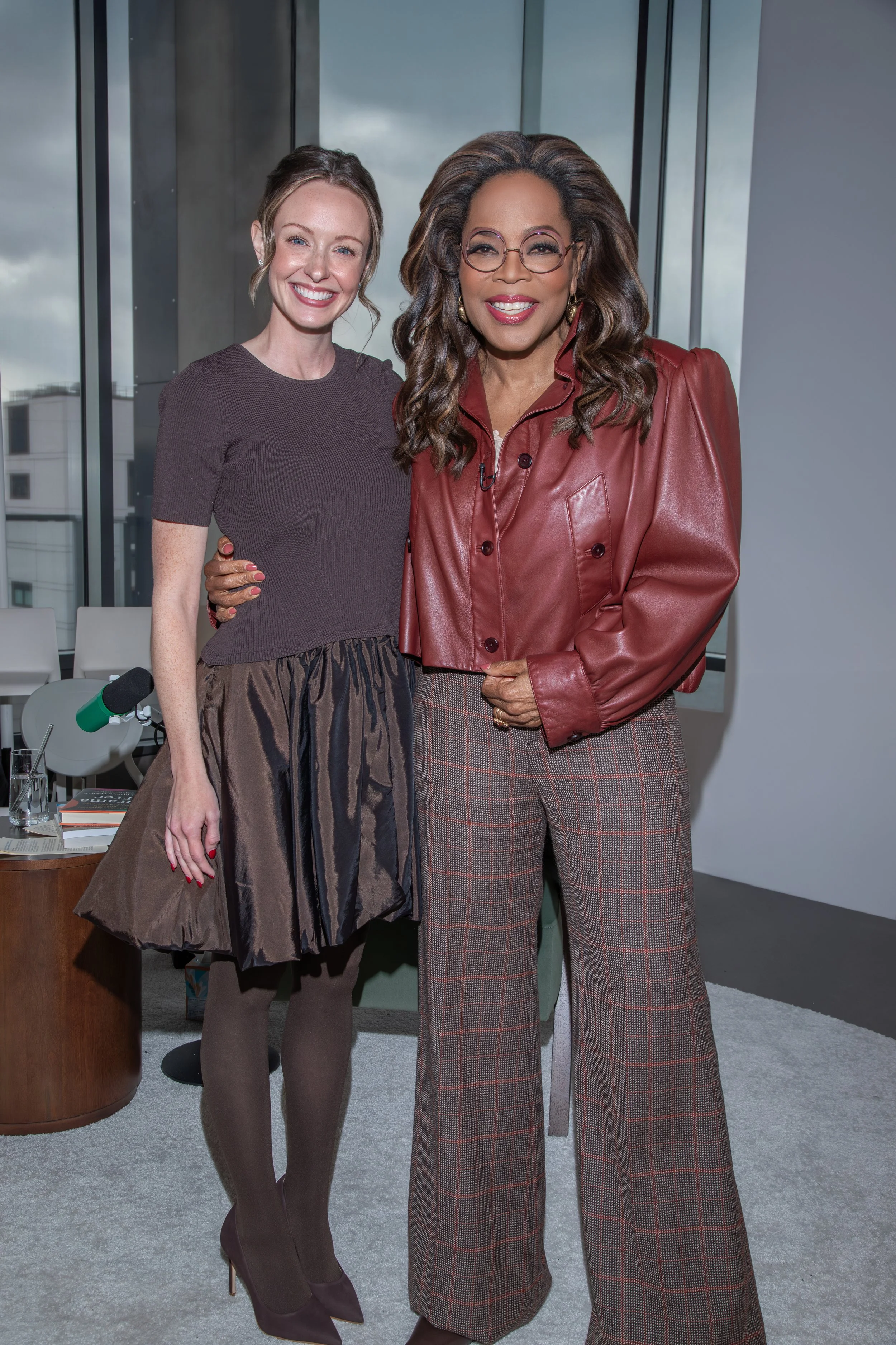 Two women posing for a photo indoors, with a cityscape visible through large windows behind them. The woman on the left has light skin, light brown hair, and is dressed in a dark brown short-sleeved top and a patterned skirt. The woman on the right has dark skin, long wavy hair, glasses, and is wearing a shiny red leather jacket and plaid pants.