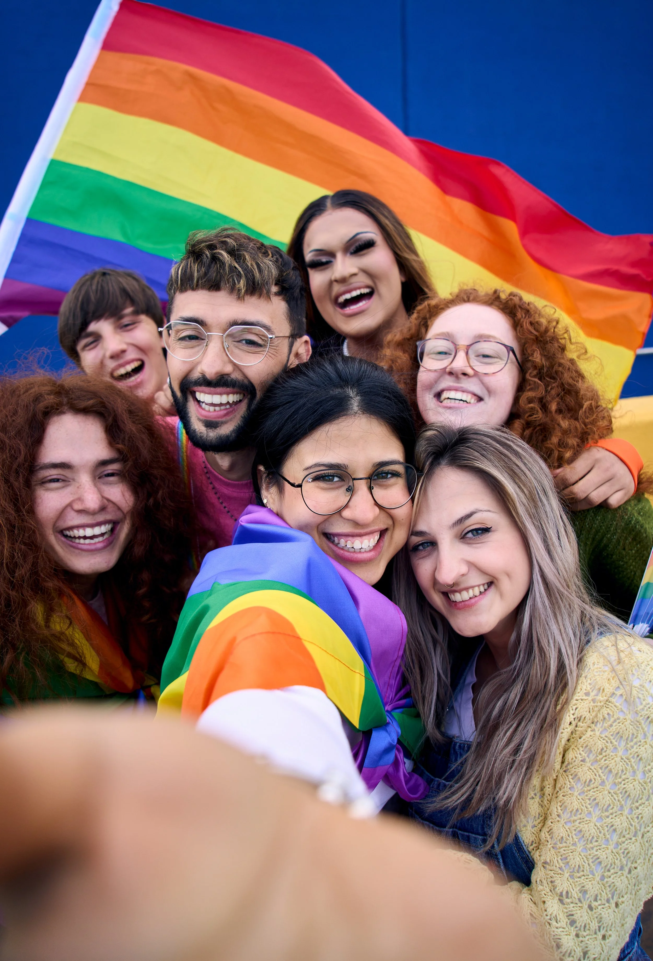 Group of diverse young adults celebrating LGBTQ+ pride, smiling and taking a selfie, with a large rainbow pride flag in the background.