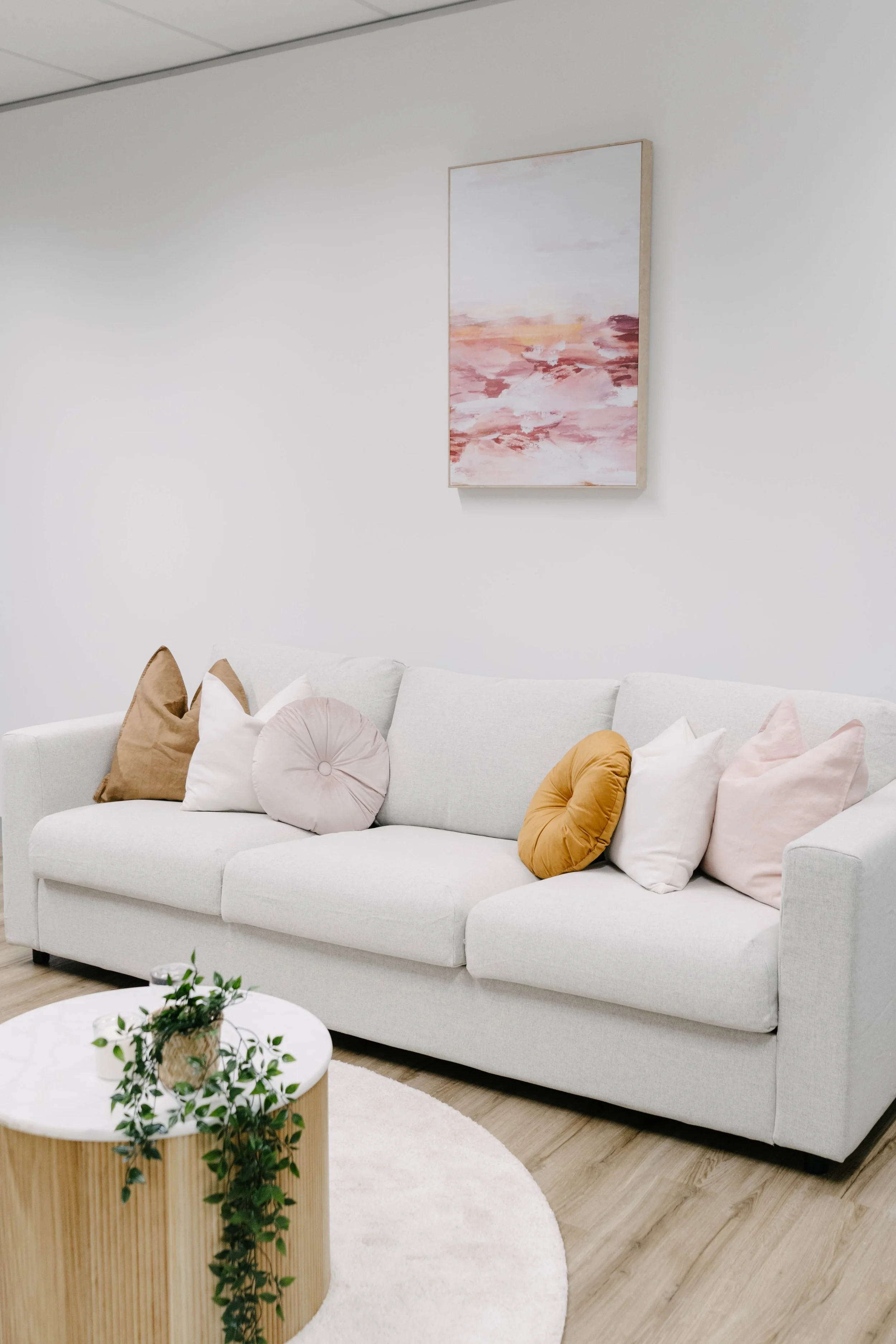 A beige sofa with a variety of pastel-colored pillows, including round and rectangle shapes, in a minimalistic room with white walls and a framed abstract pink and red painting hanging above.