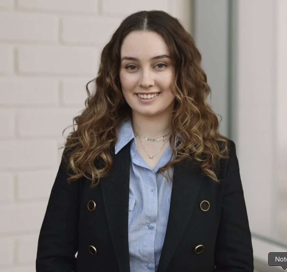 A young woman with long, curly brown hair smiling in front of a white brick wall, dressed in a black blazer and blue shirt, wearing layered necklaces.