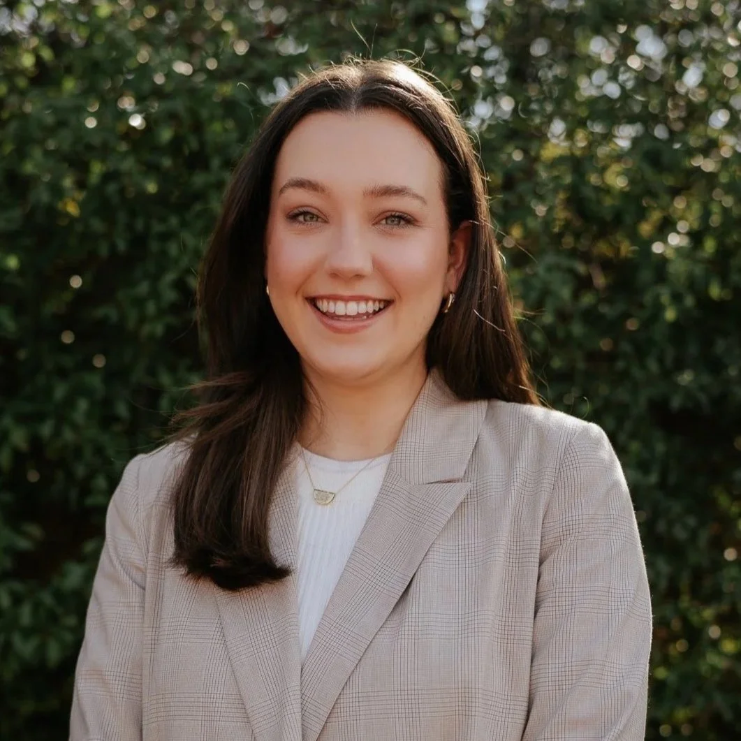 A woman with long dark hair smiling, wearing a beige blazer with a plaid pattern and a white top, standing outdoors in front of green bushes.