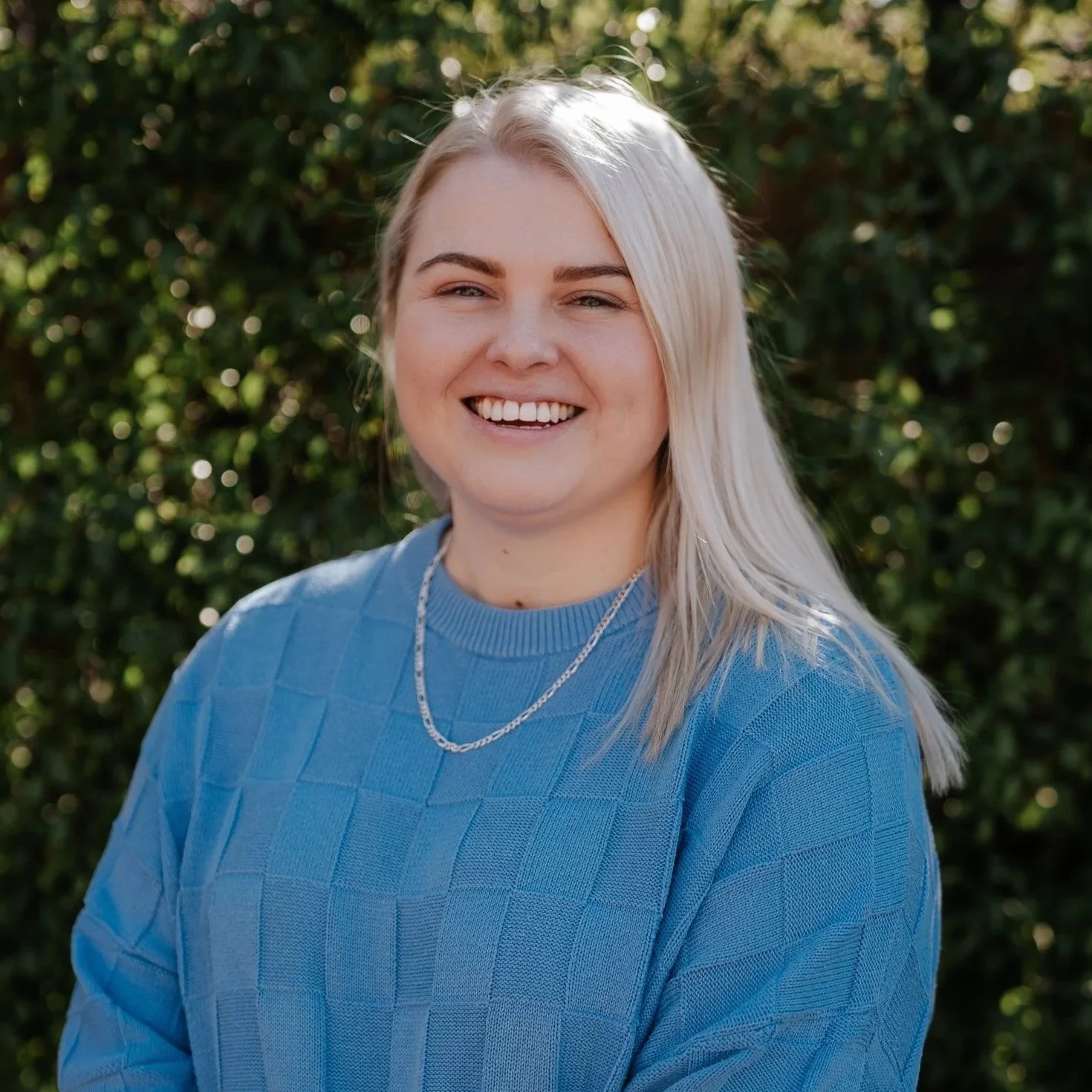 Close-up of a smiling woman with long blonde hair wearing a blue sweater and a silver necklace, standing outdoors with green foliage background.