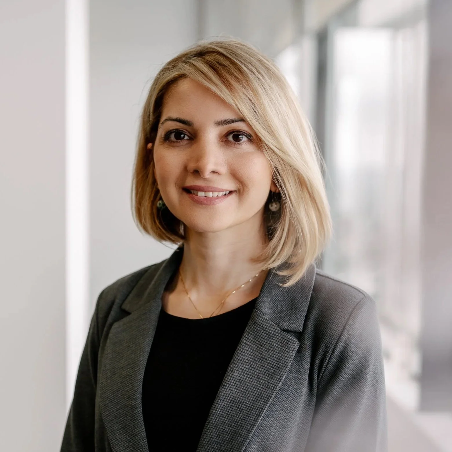 Smiling woman with short blonde hair wearing a grey blazer and black top, standing in a bright modern interior space with large windows.